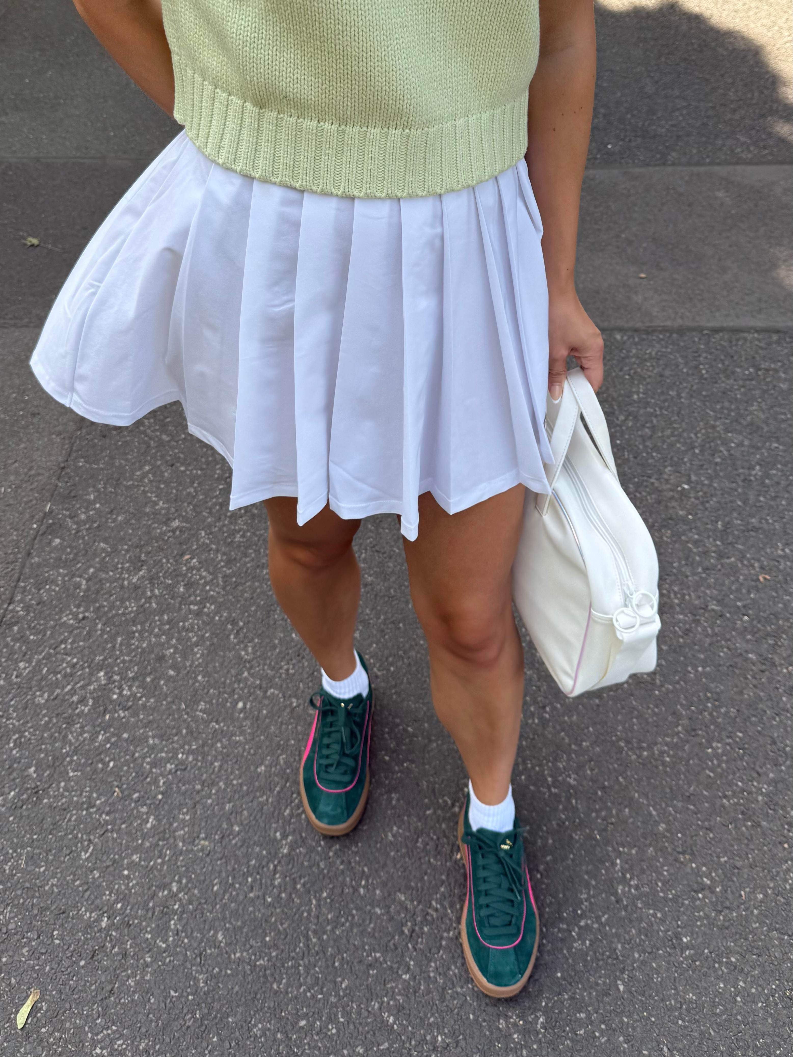 Frau in weißem Tennisrock und Retro-Sneakern steht entspannt