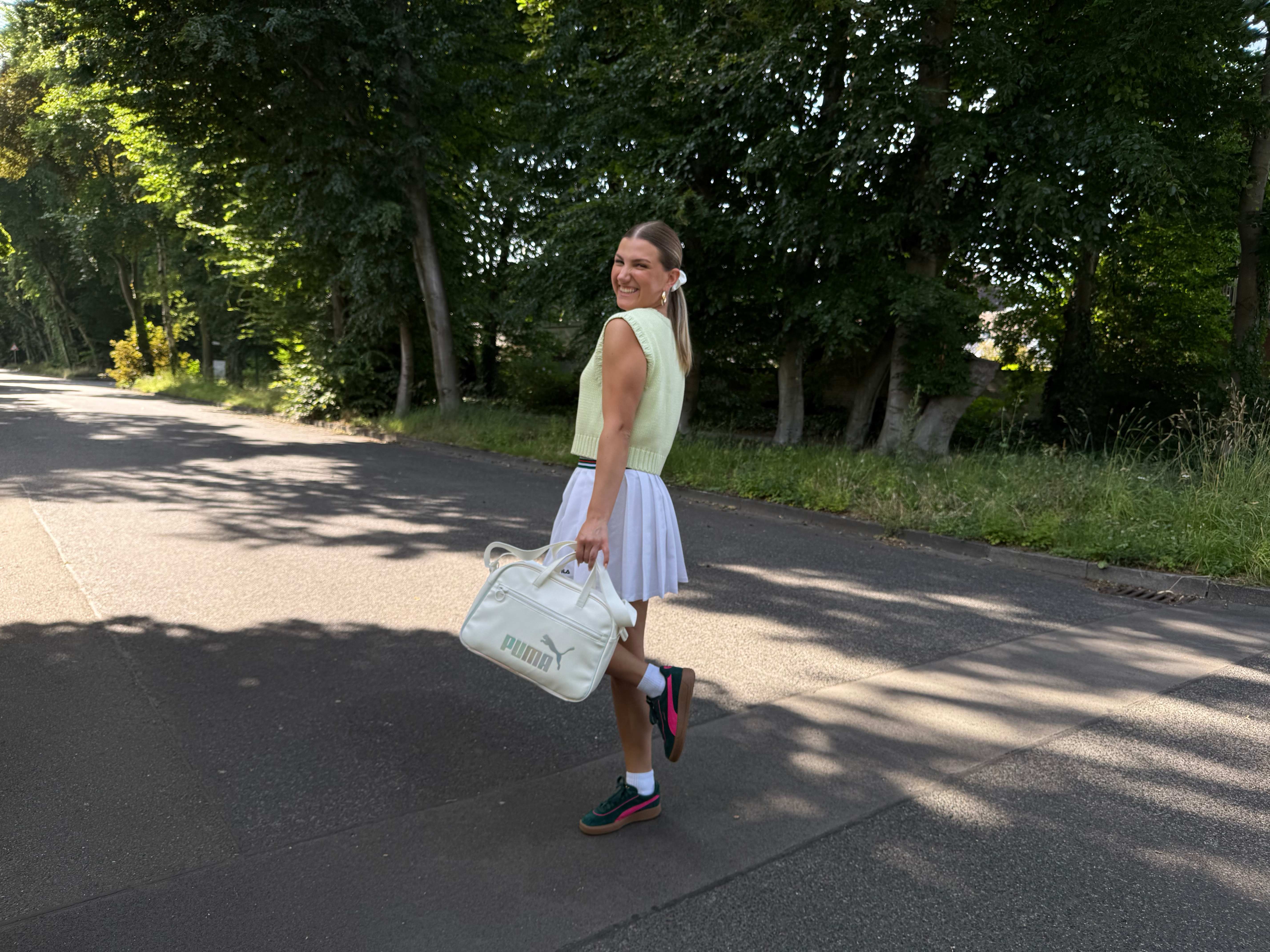 Frau in gelb-weißem Tennis-Outfit steht auf Straße, trägt Puma-Tasche