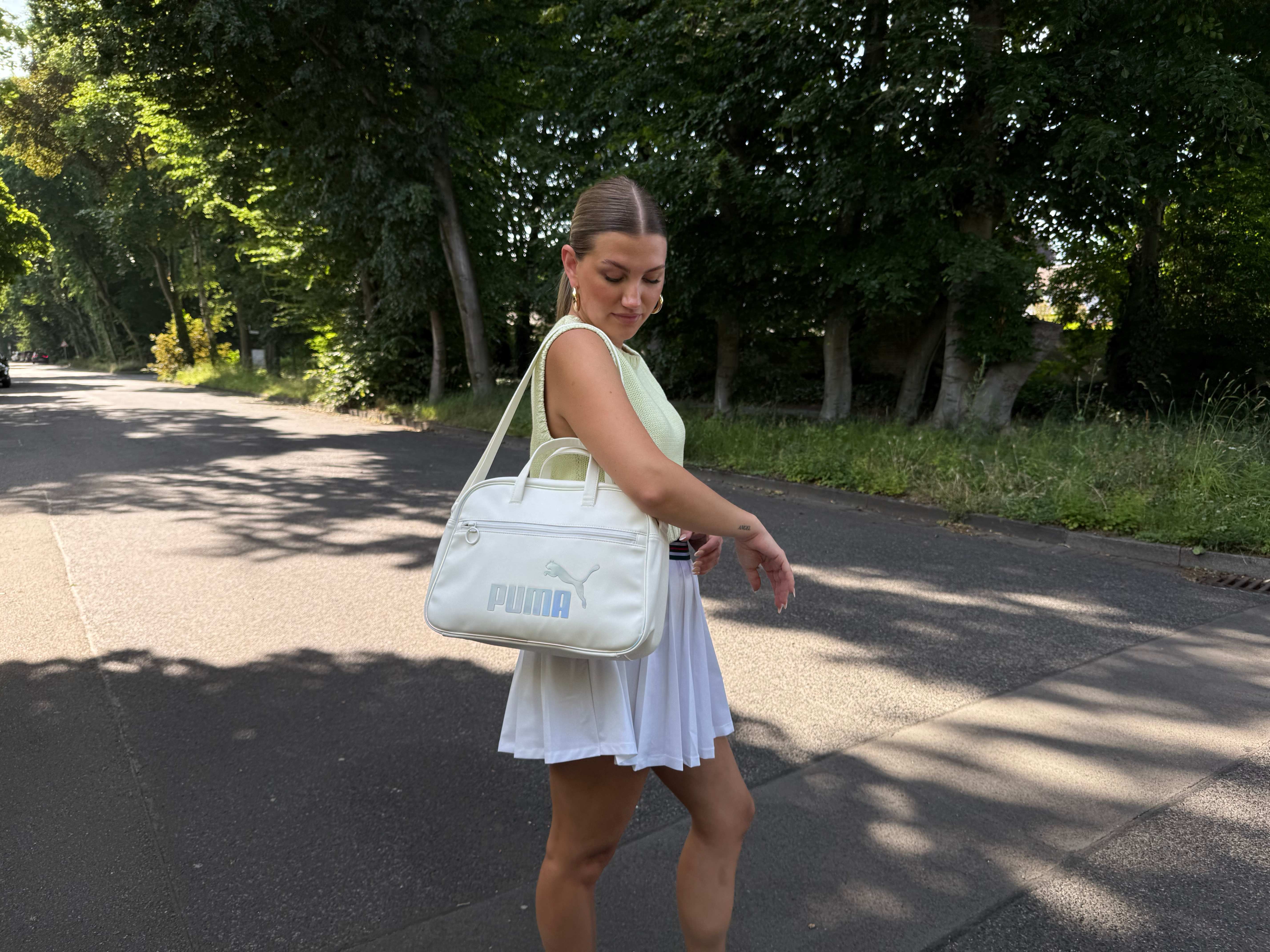 Frau in gelb-weißem Tennis-Outfit, trägt Puma-Tasche am Arm, posiert vor Waldstück