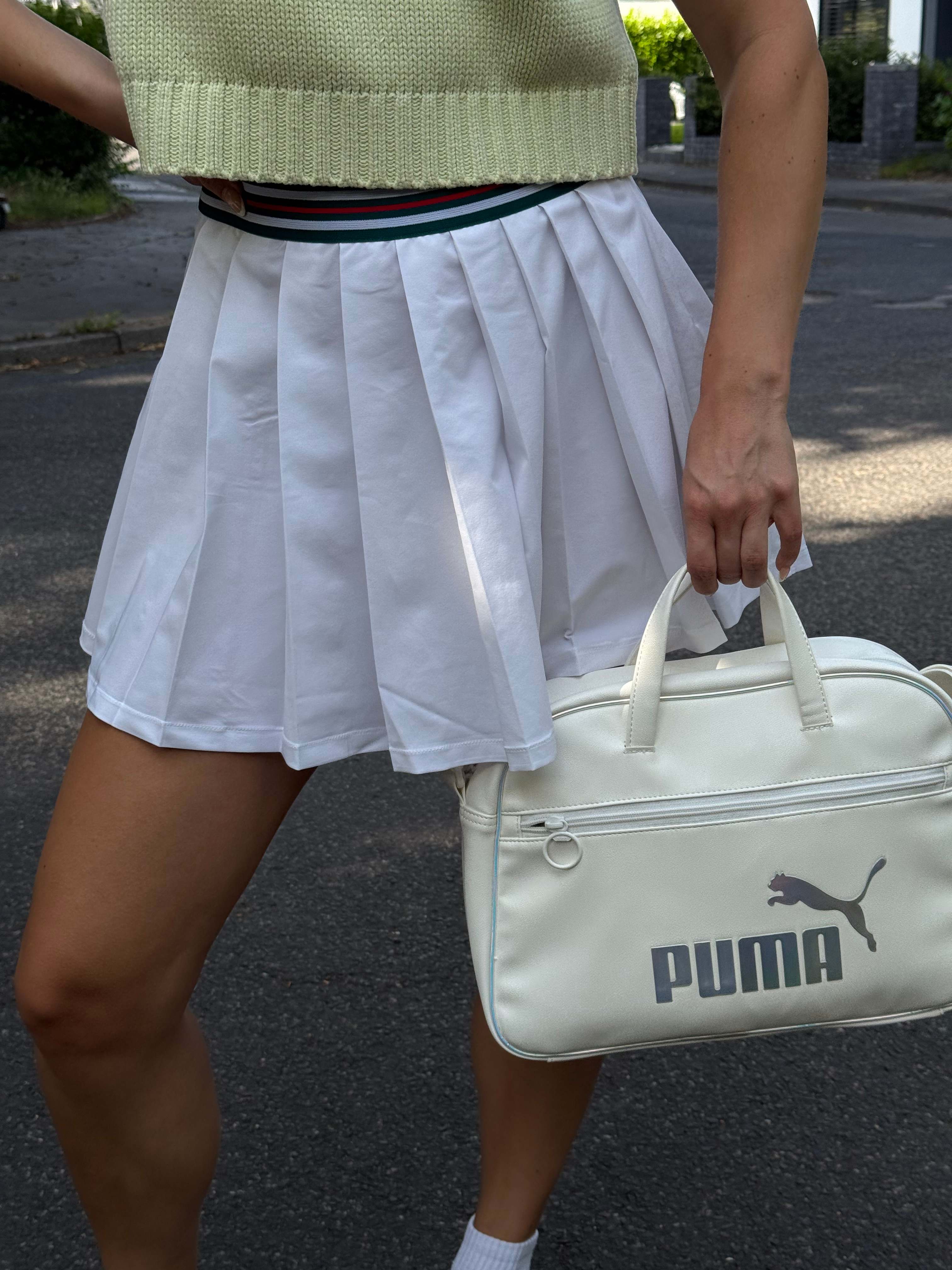 Sportliche Frau präsentiert Outfit mit Tennis-Rock und beiger Puma-Handtasche