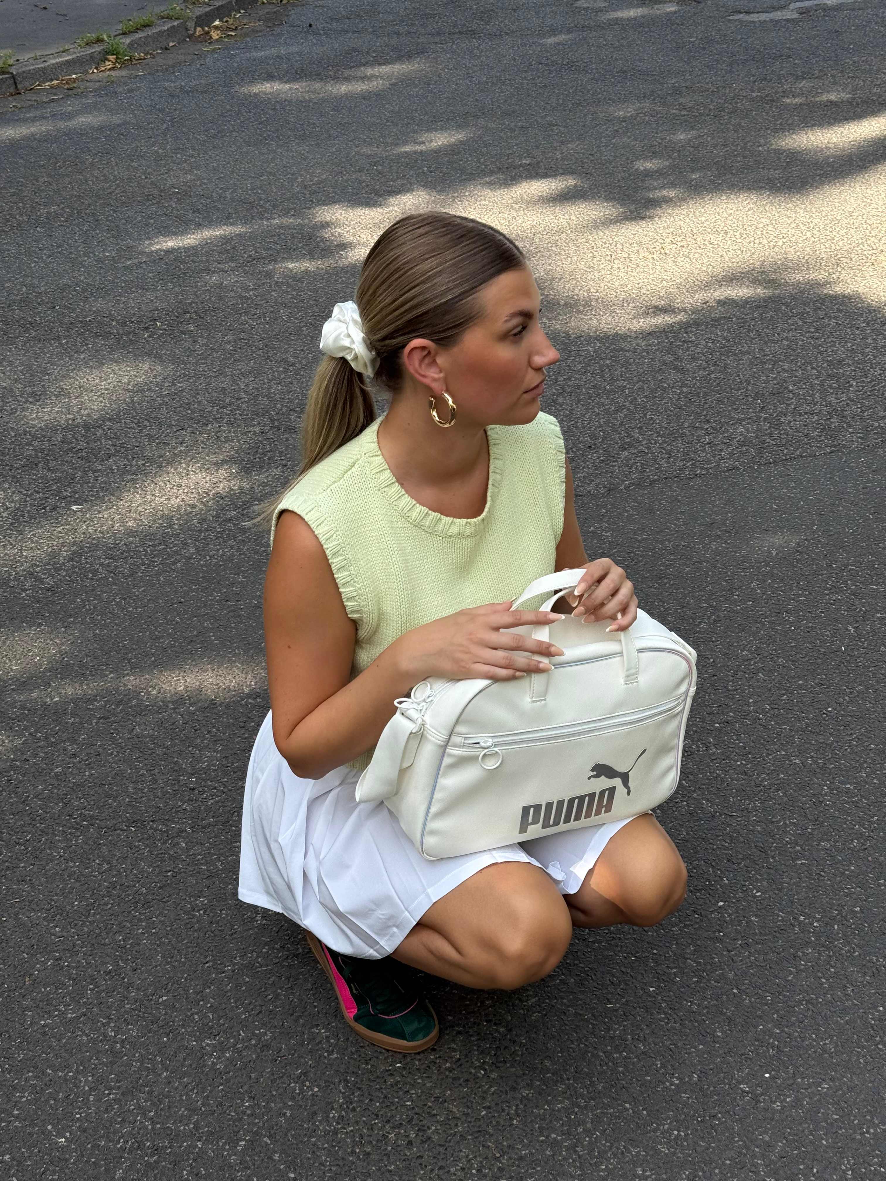 Frau in Tennis-Outfit hockt auf Asphaltboden, hält Puma-Tasche auf den Knien