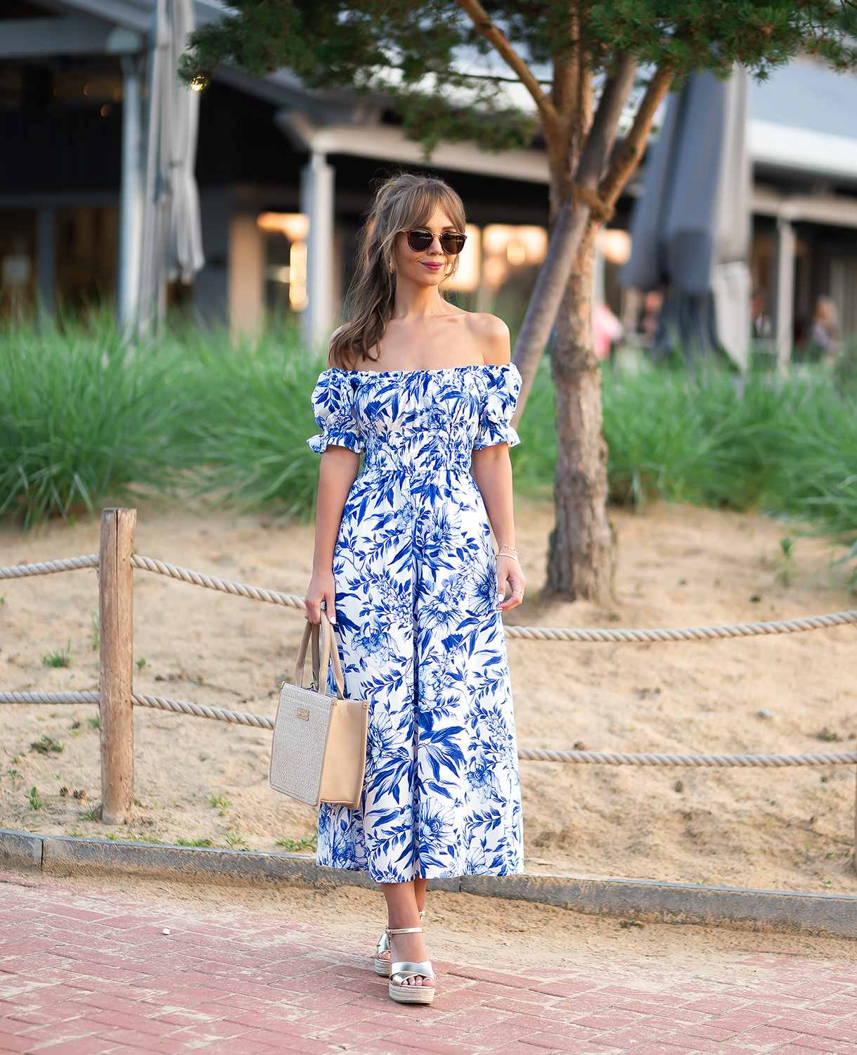 Frau trägt sommerliches Strandoutfit, bestehend aus blauem schulterfreiem Kleid mit Sandalen und Handtasche