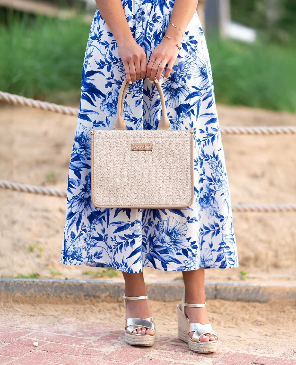 Frau trägt Strandoutfit aus blauem Sommerkleid und Sandalen im Metallic-Look, kombiniert mit Handtasche