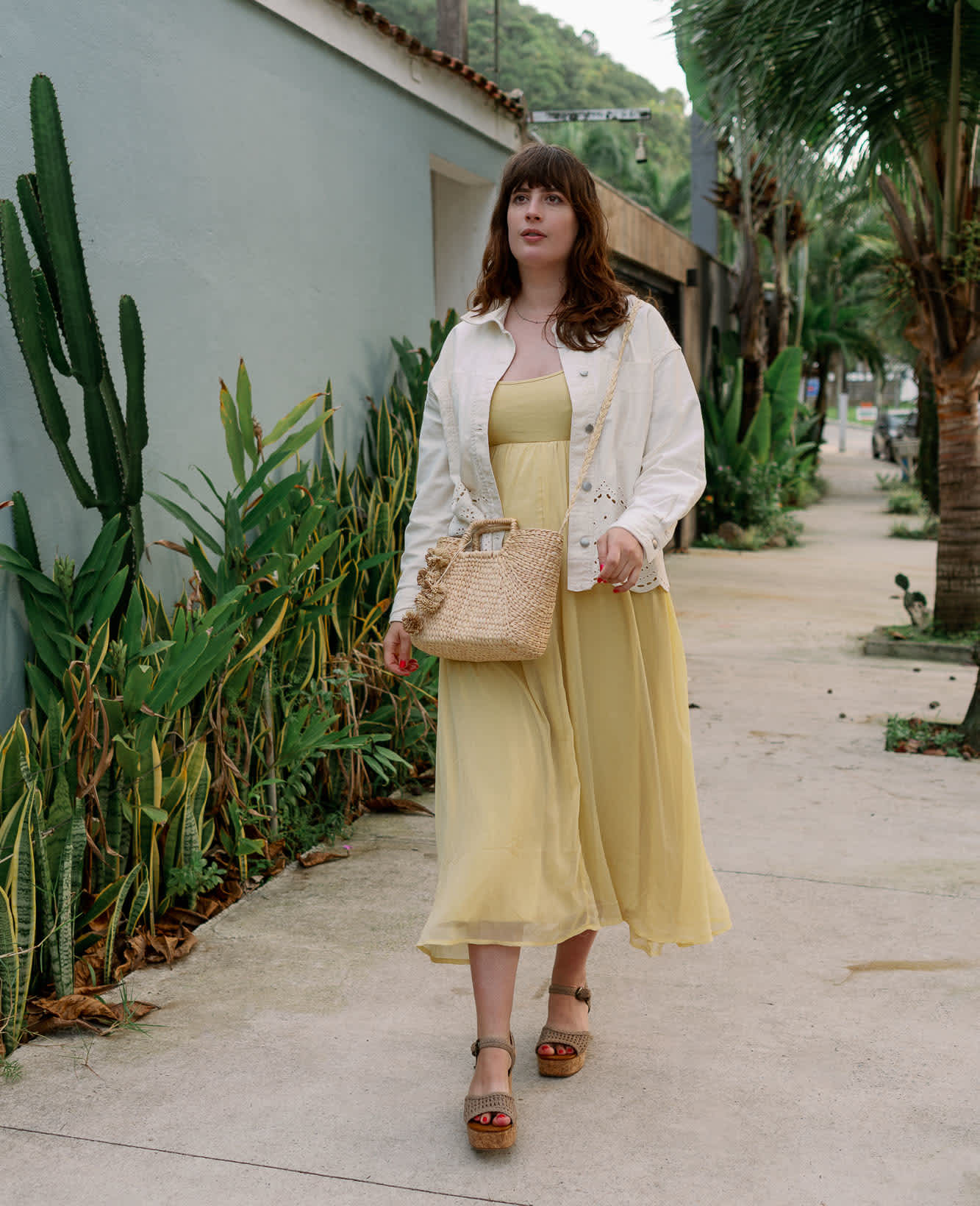 Frau trägt ein sommerliches gelbes Kleid, eine weiße Jacke, eine Boho-Handtasche und dazu passende Keilsandaletten