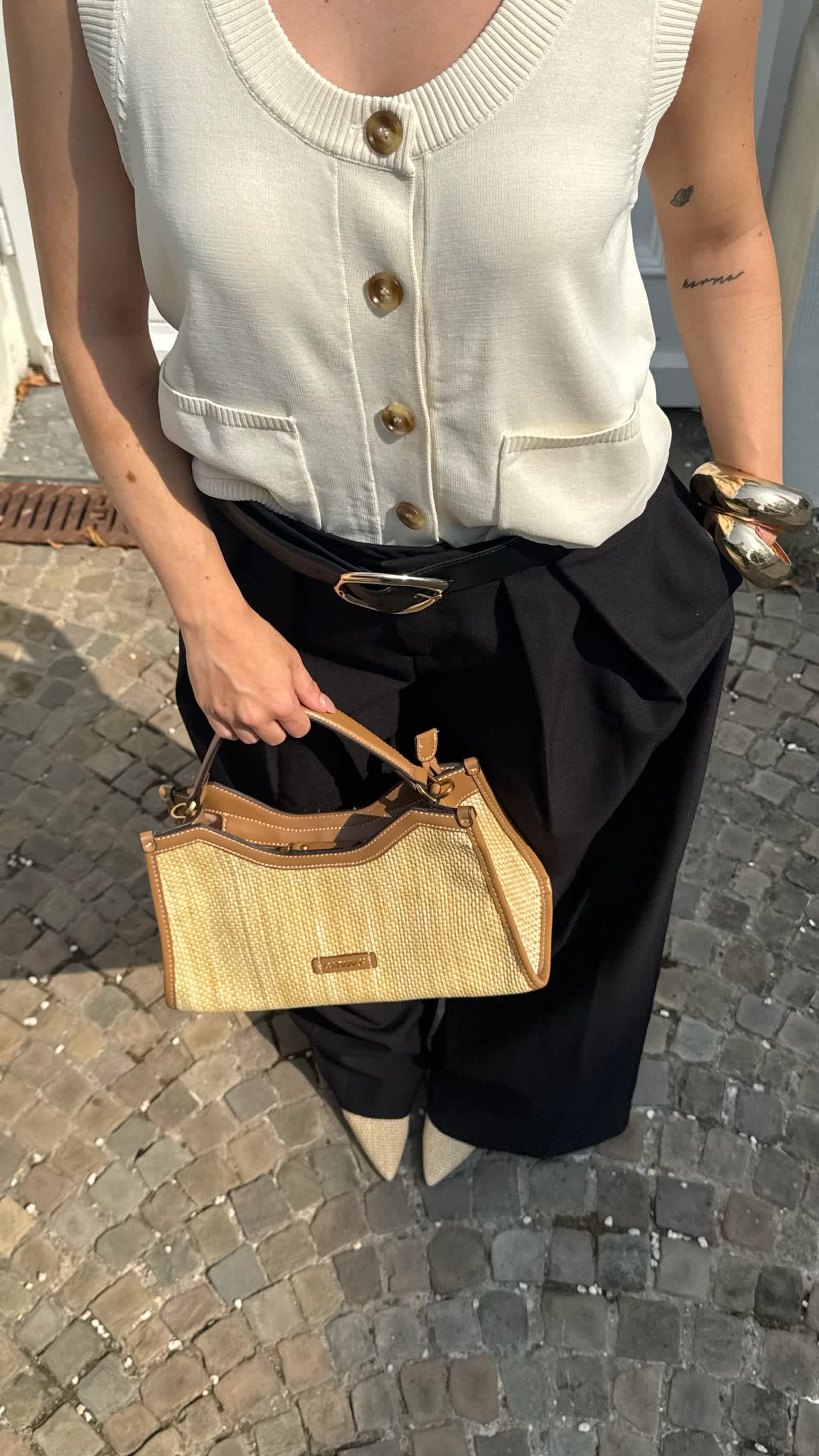 Frau zeigt Details ihres Old Money Outfits: goldene Gürtelschnalle, goldene Armreifen und Handtasche in Bastoptik