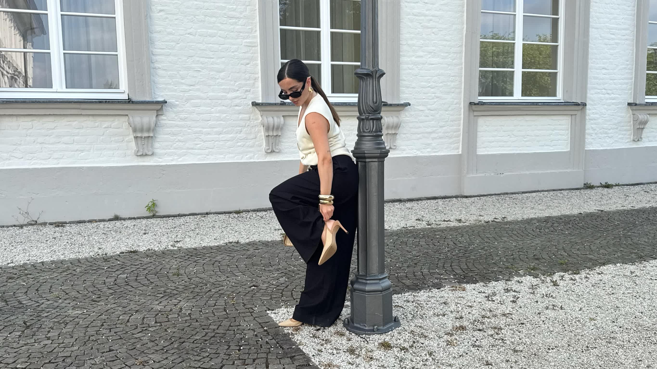 Frau präsentiert ihr Old Money Fashion Outfit mit weiter schwarzer Anzughose, ärmelloser Weste und cremefarbenen Schuhen