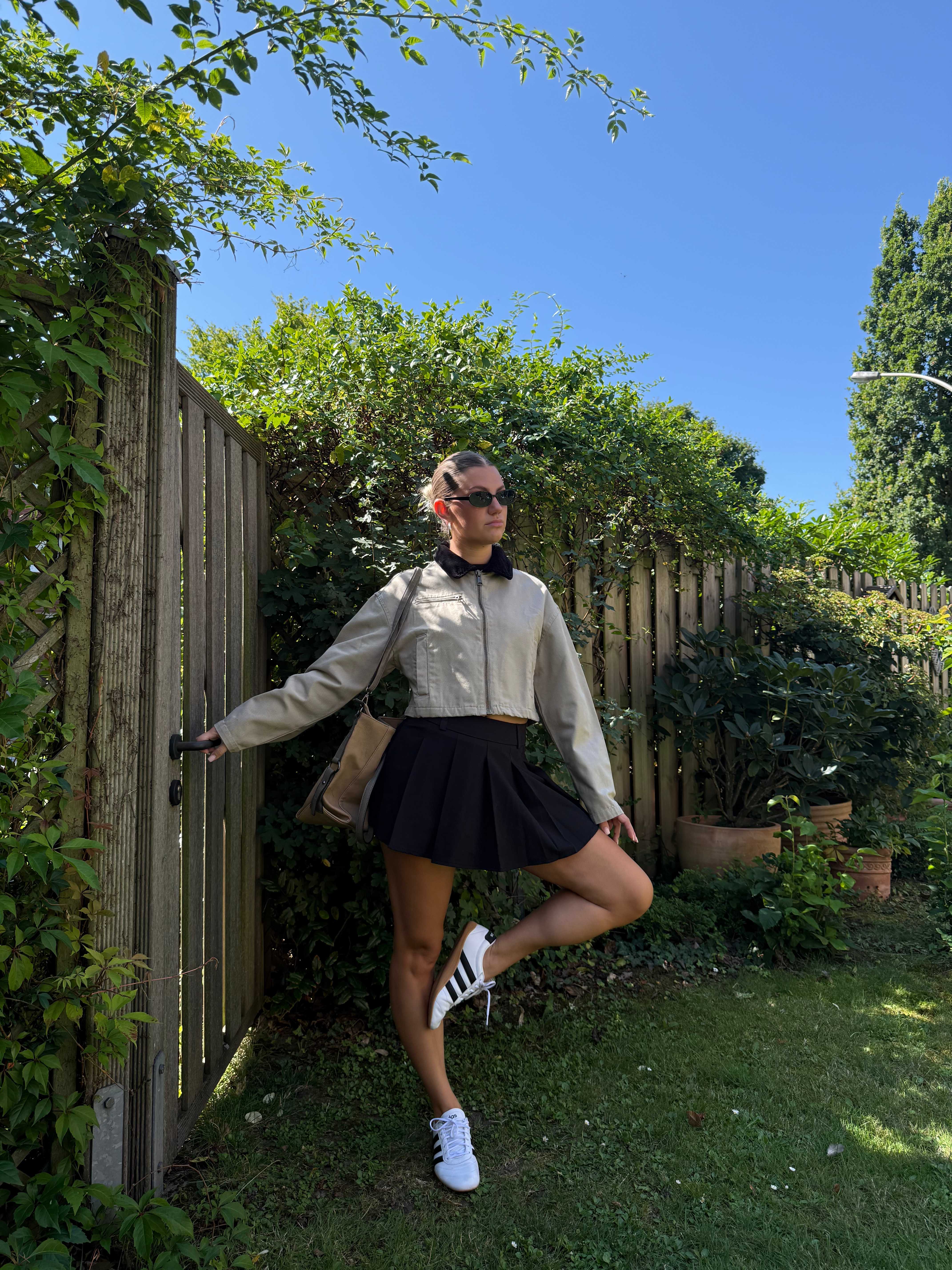 Frau in grauem Top und schwarzem Rock steht im Garten, mit der Hand an einem Holztor