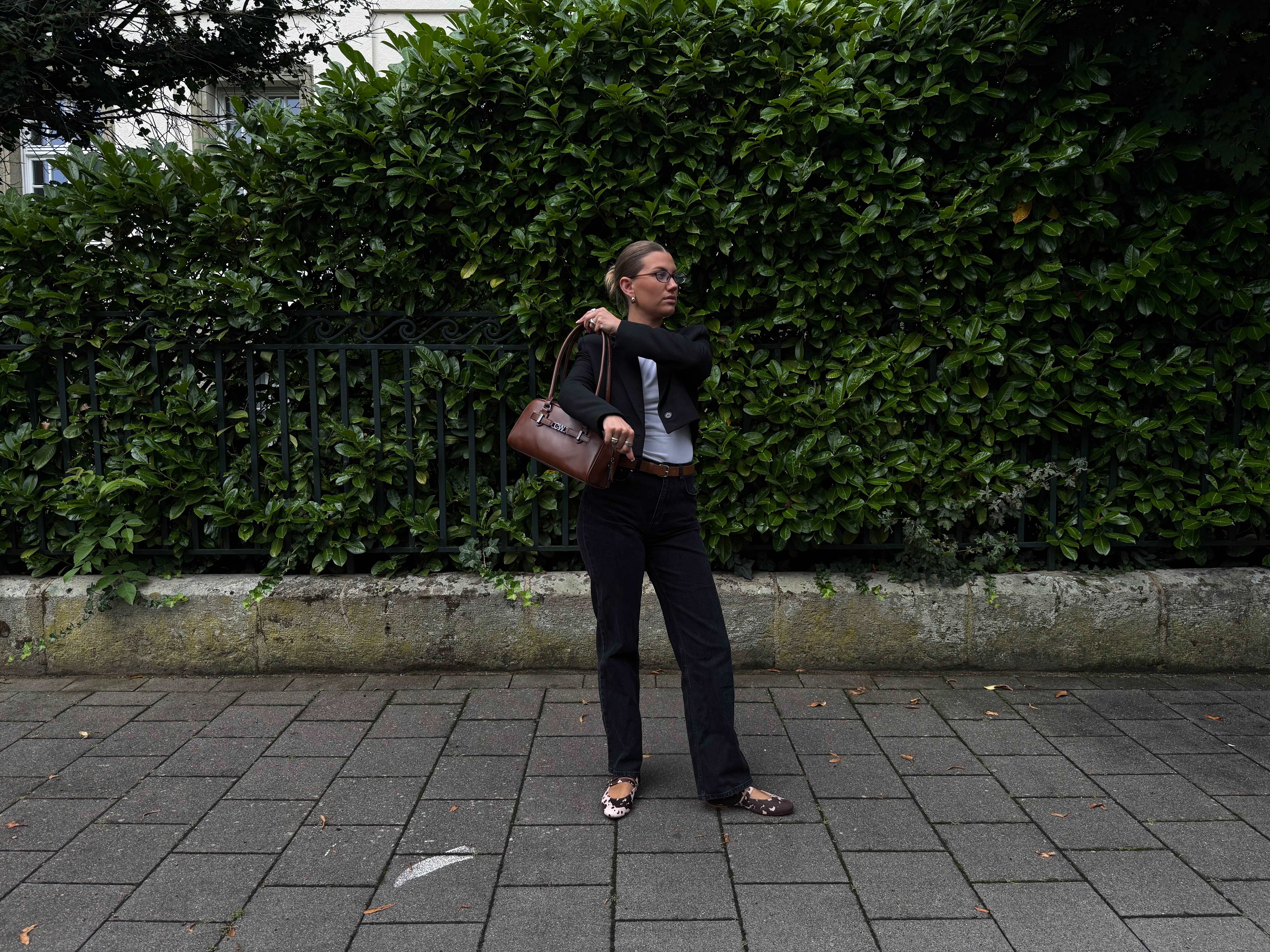 Frau steht vor Hecke und schwingt sich braune Handtasche über die Schulter