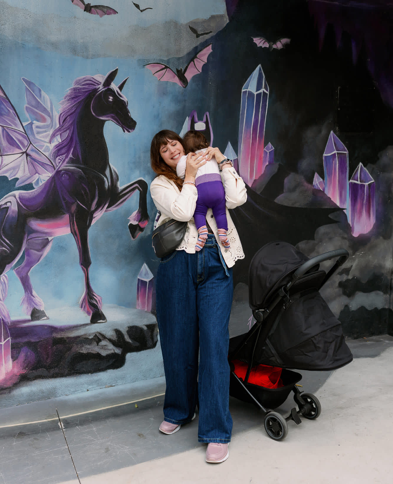 Frau mit Baby und Kinderwagen steht vor Wand mit lila Einhorn-Painting