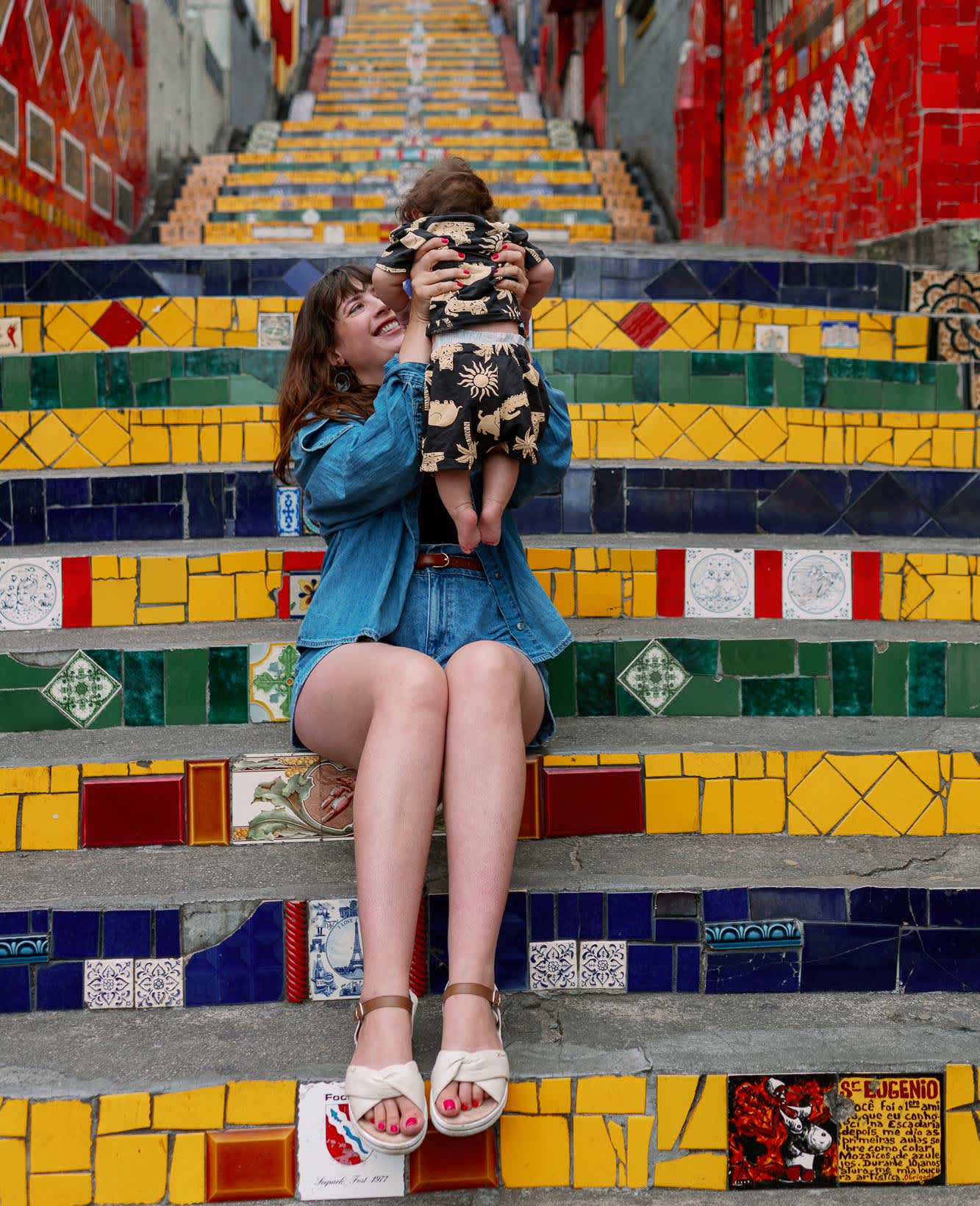 Frau hält Baby in die Luft, während sie auf bunt gefliesten Treppenstufen sitzt