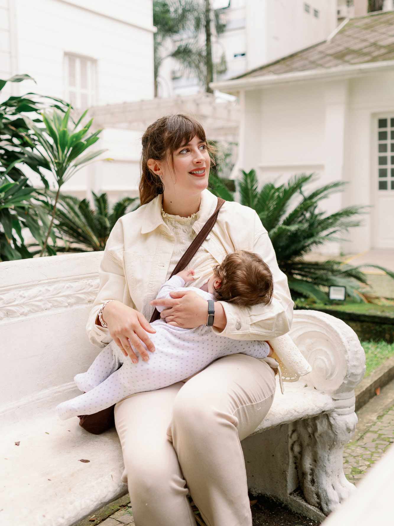 Lächelnde Frau in weißem Outfit sitzt auf Steinbank, hält Baby im Arm