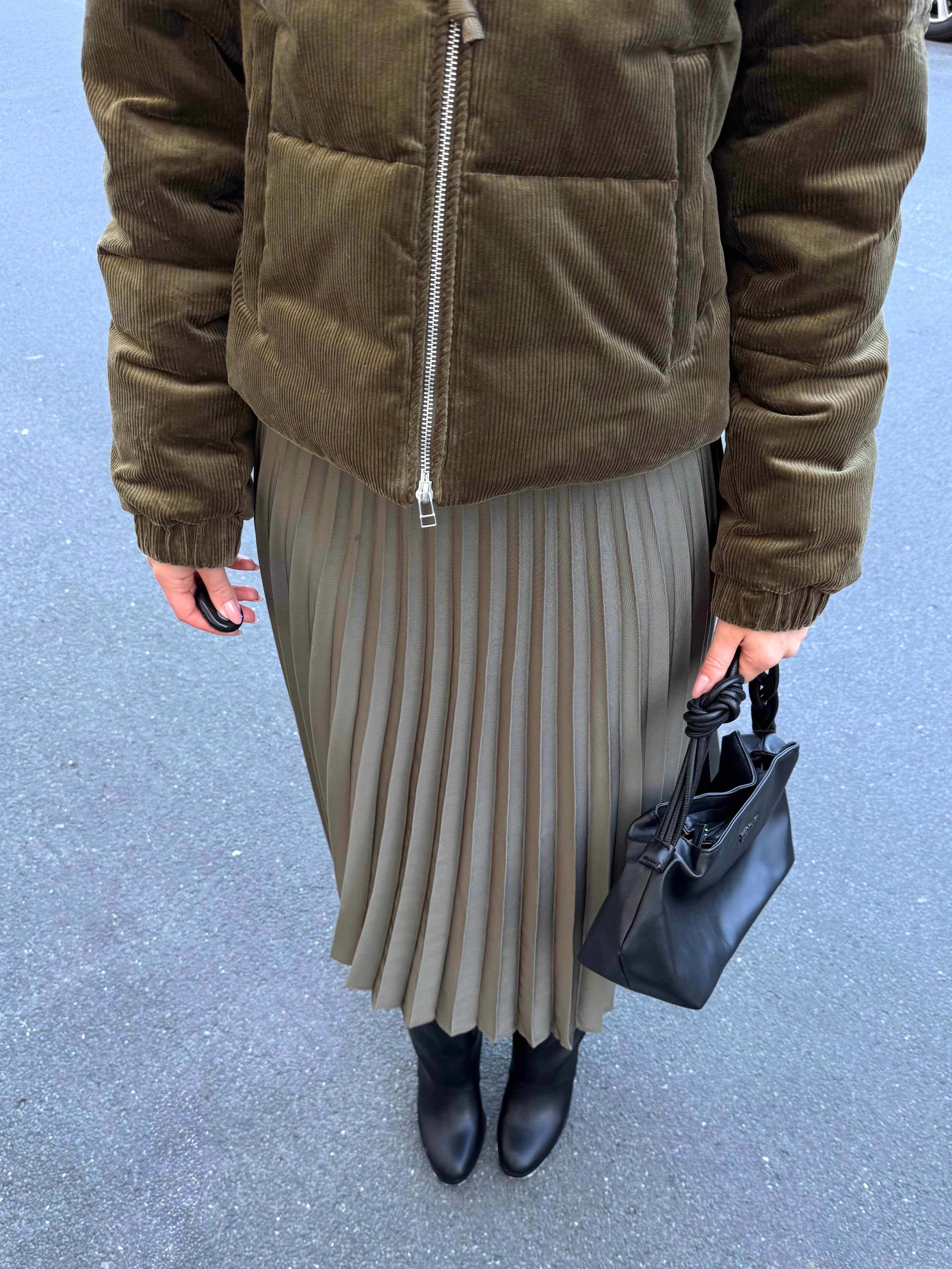 Frau in grüner Winterjacke, Faltenrock und Underknee Boots, steht entspannt