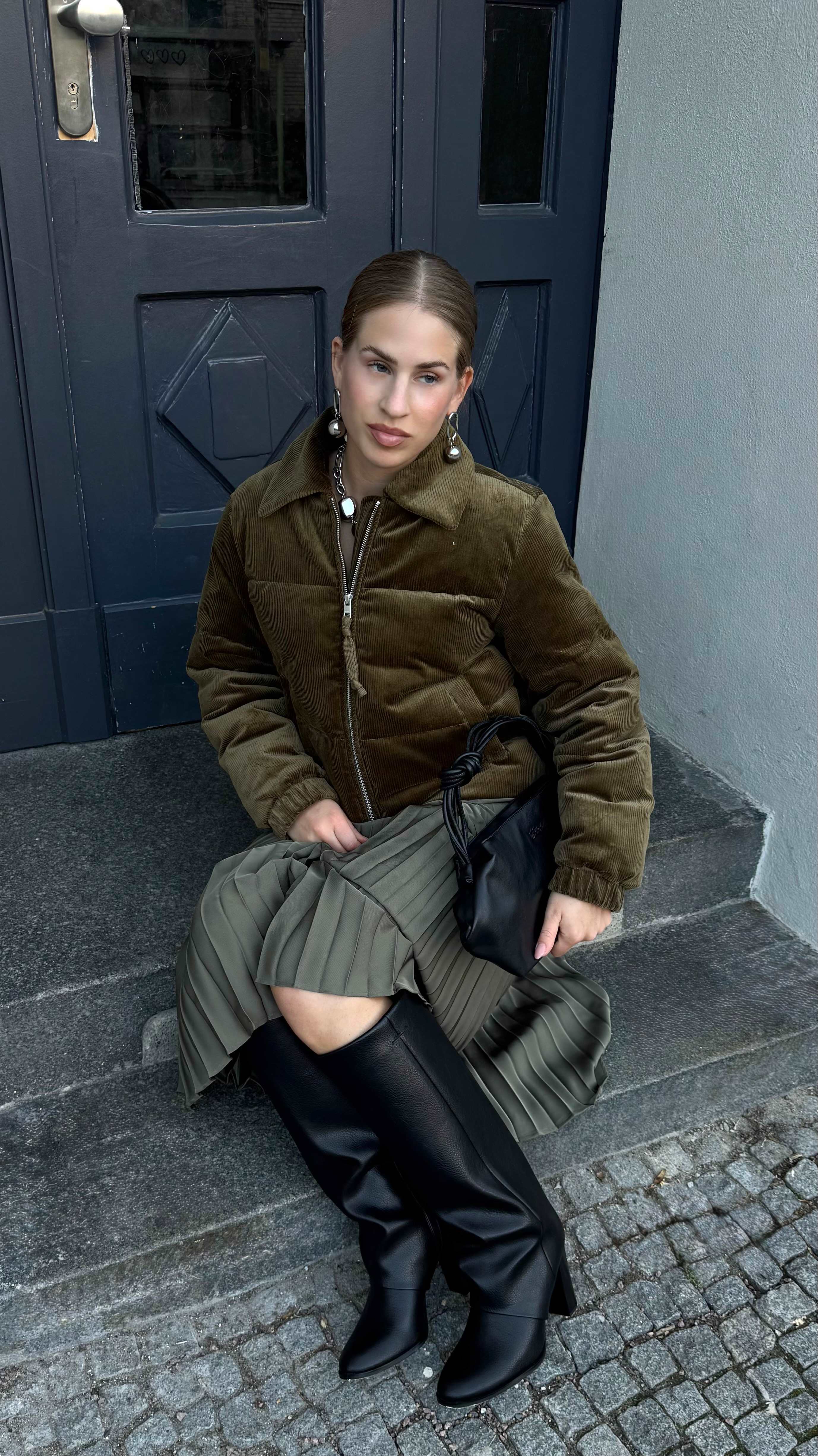 Frau in dunkelgrüner Jacke, mit salbeifarbenem Rock und Underknee Boots, sitzt auf Steintreppe