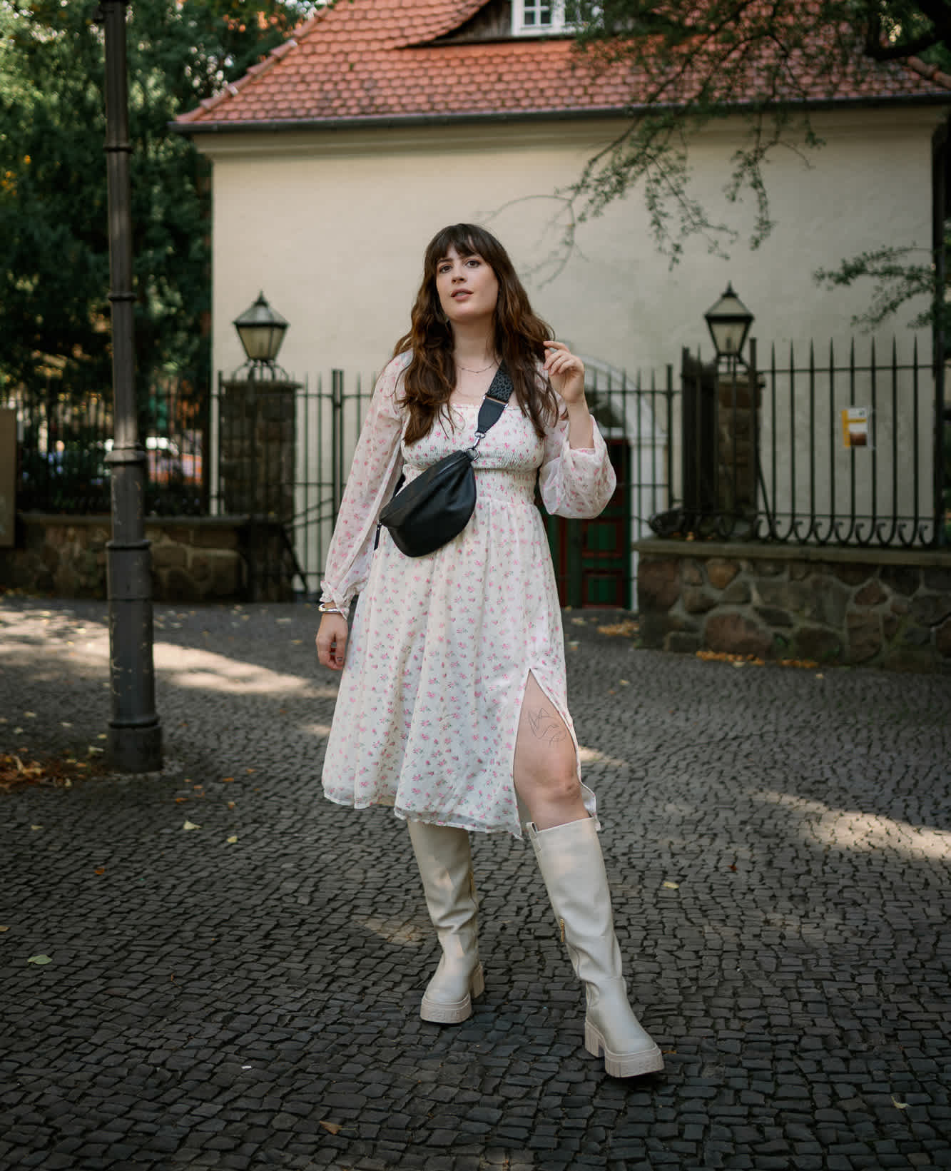 Frau in weißem Spitzenkleid mit Underknee-Boots und schwarzer Brusttasche