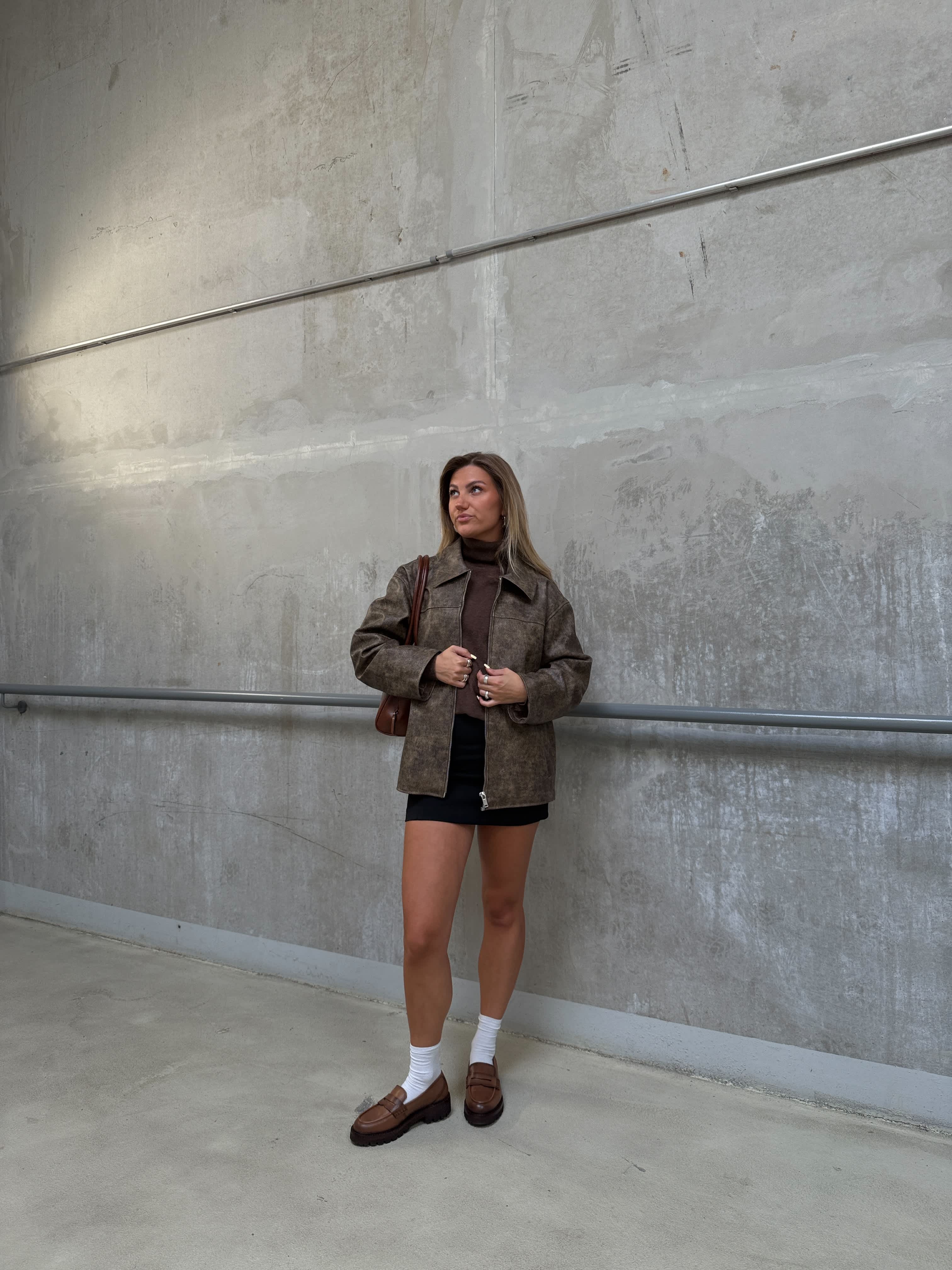 Frau in brauner Jacke, mit schwarzen Pants und braunen Loafern, steht vor Betonwand