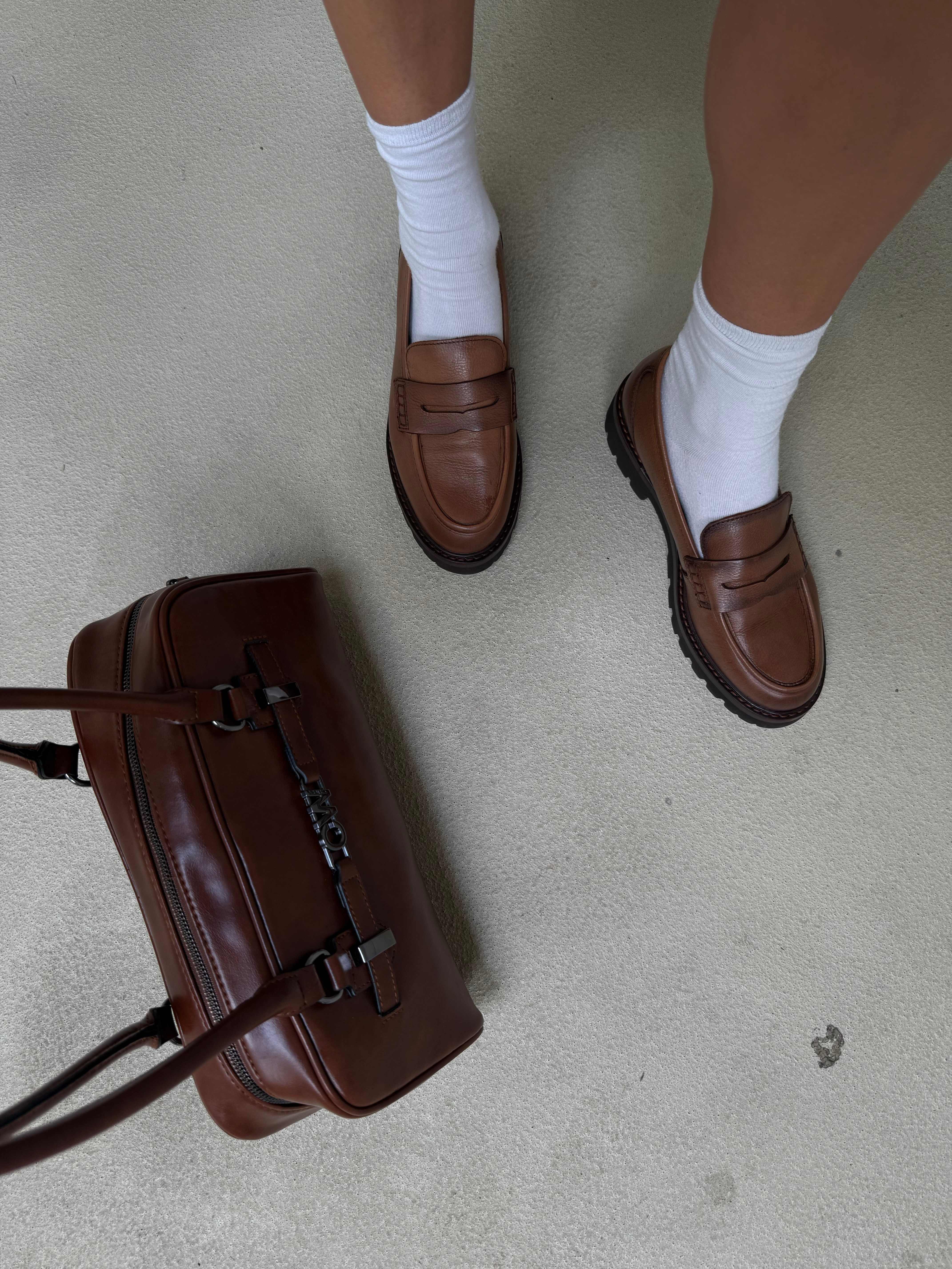 Top-Down-Ansicht einer Person, die in braunen Loafern neben einer passenden Handtasche steht