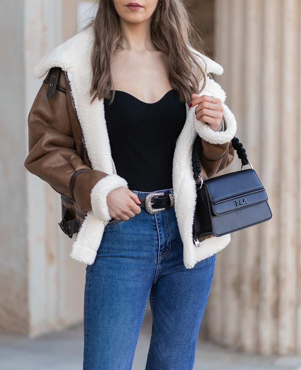 Frau mit brauner Jacke, schwarzem Top, Jeanshose und schwarzer Handtasche steht entspannt