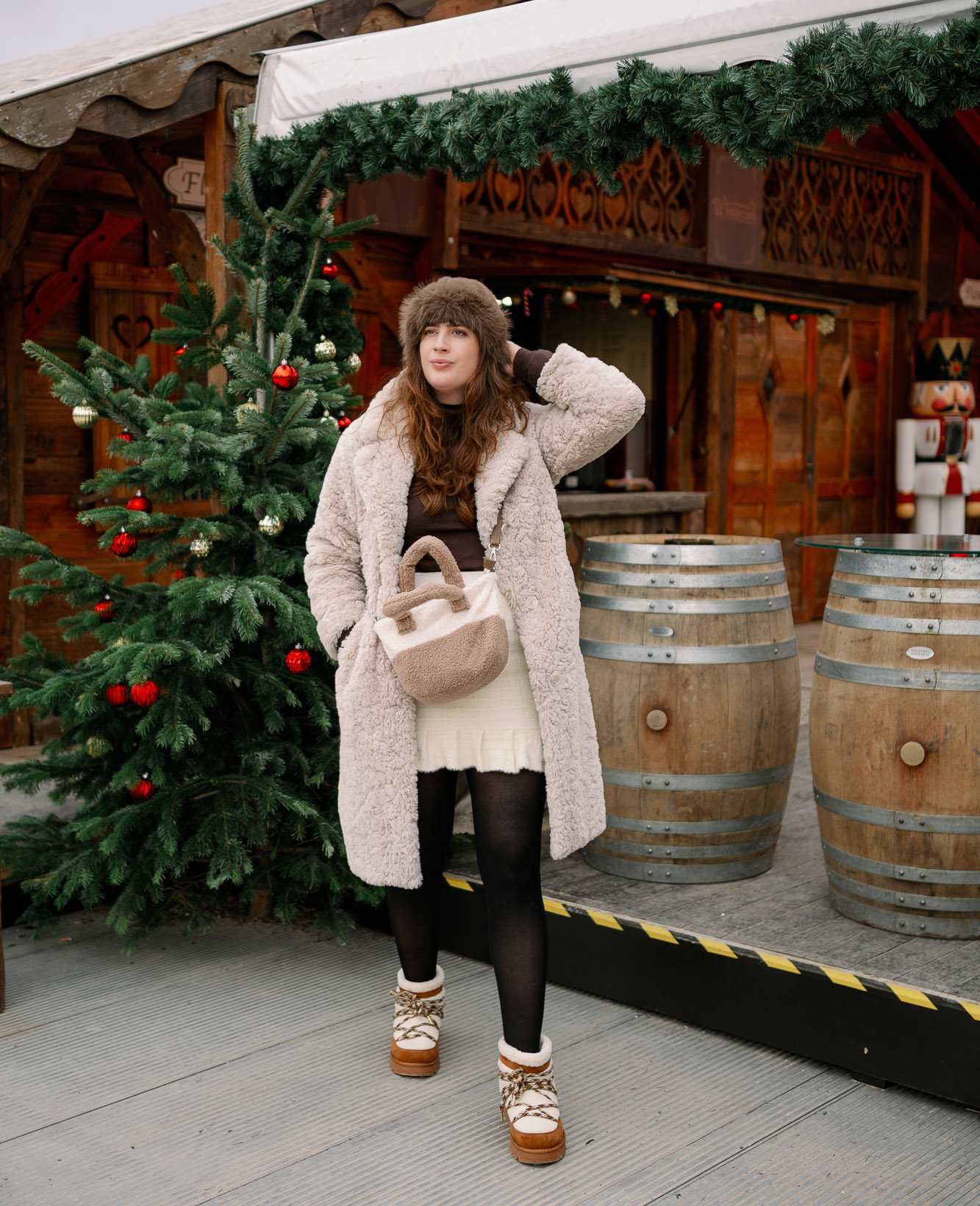 Frau in flauschigem Outfit, mit Mantel, Boots und Tasche in Plüsch, steht vor Weihnachtsmarkt-Kulisse