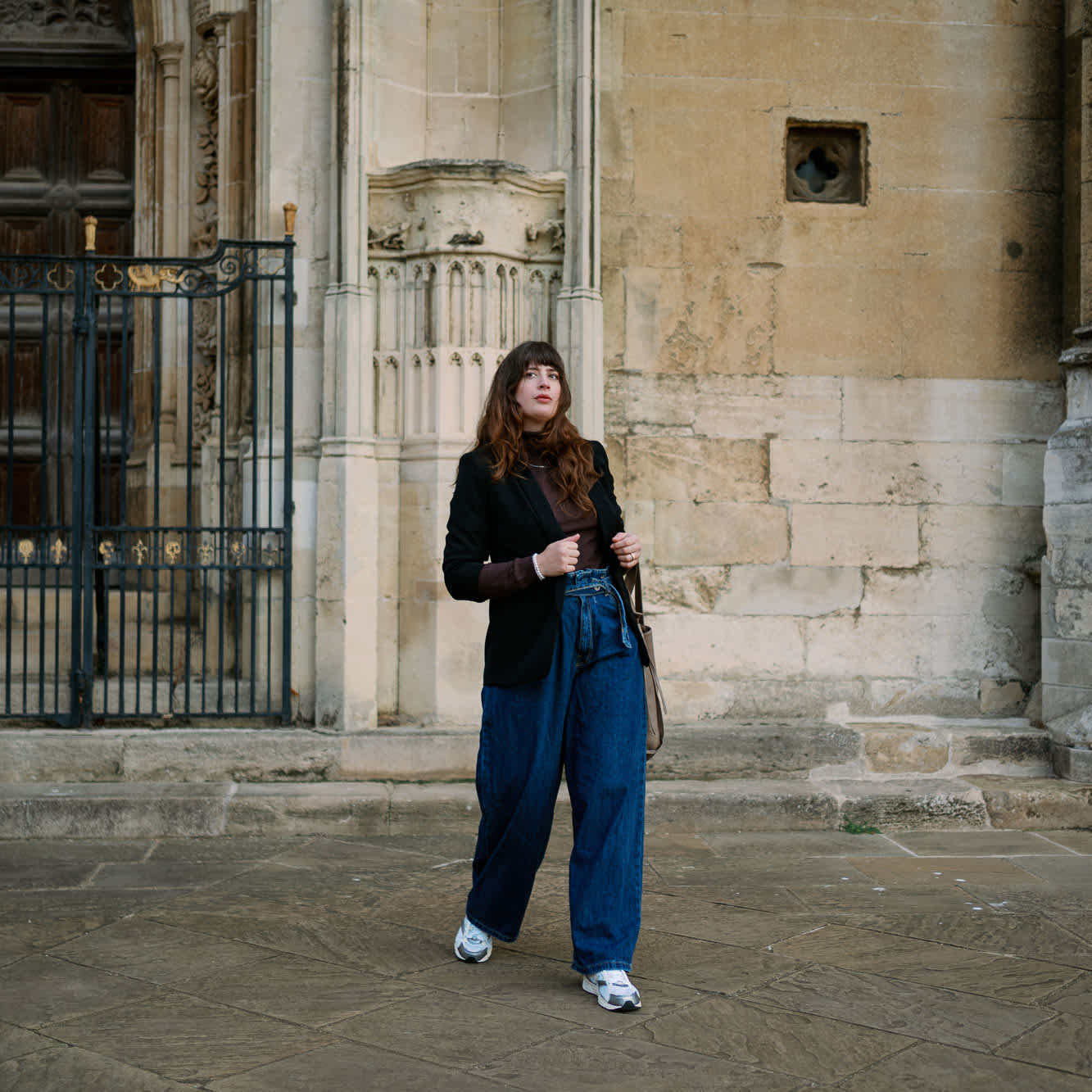 Brunette Frau trägt Outfit im Normcore-Style, steht vor Steinfassade eines Gebäudes