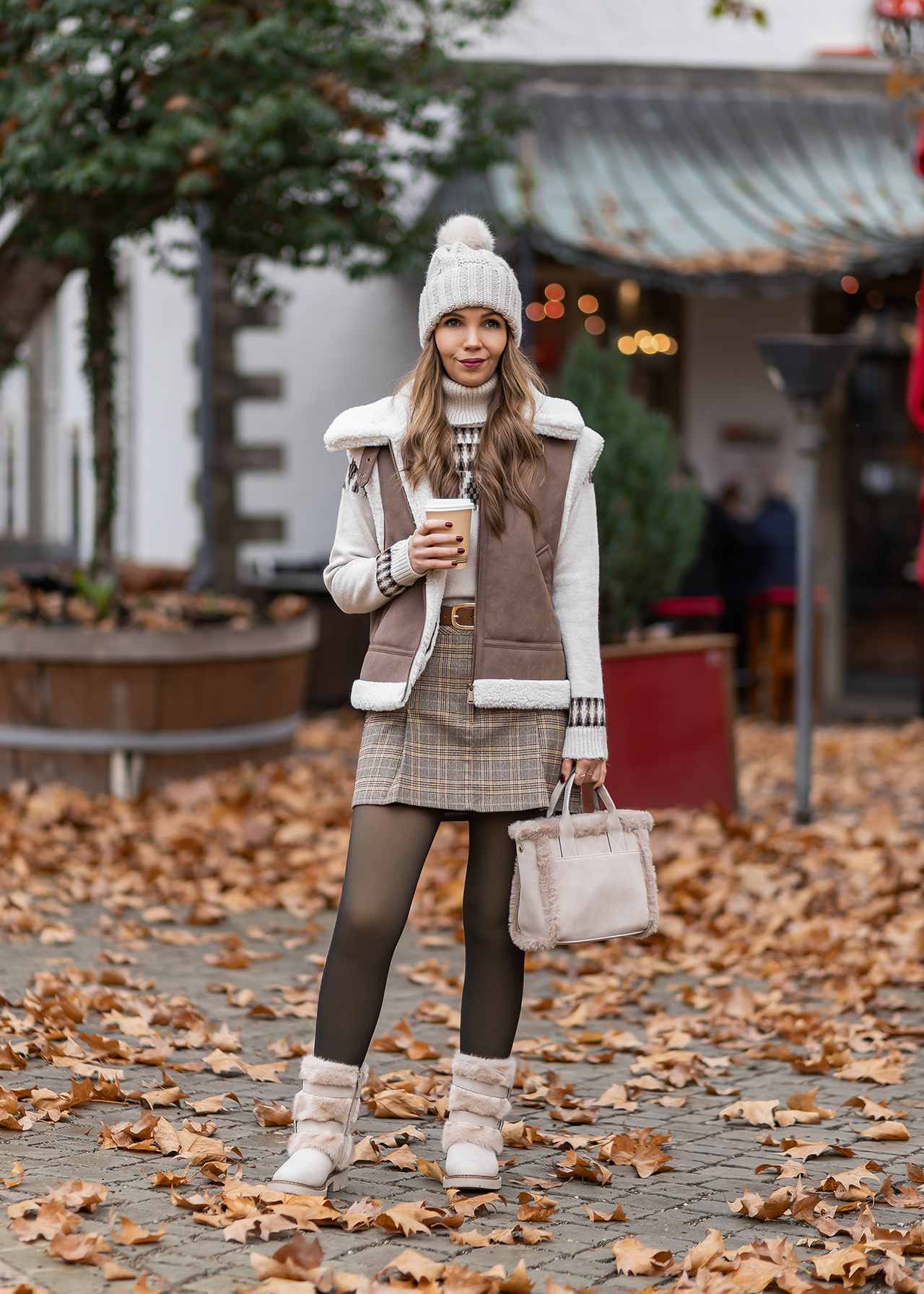 Frau in Winter-Outfit mit beigefarbener Strickmütze, braun-weiß kariertem Rock, weißem Pullover, Shearling-Weste und cremefarbenen, pelzbesetzten Winterstiefeln.