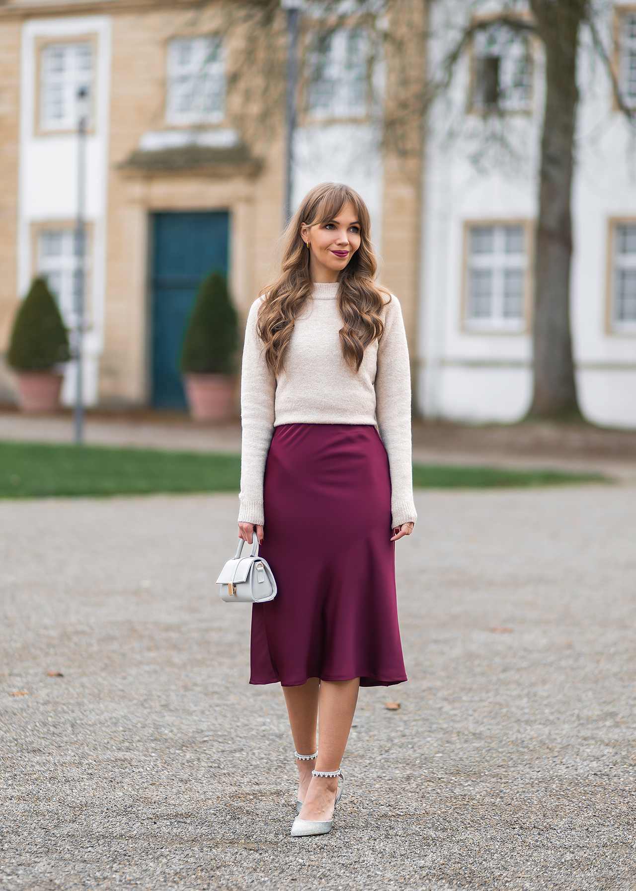 Frau in beigem Strickpullover, weinrotem Satinrock, mit kleiner weißer Handtasche und weißen Riemchensandalen vor einem hellen Gebäude.