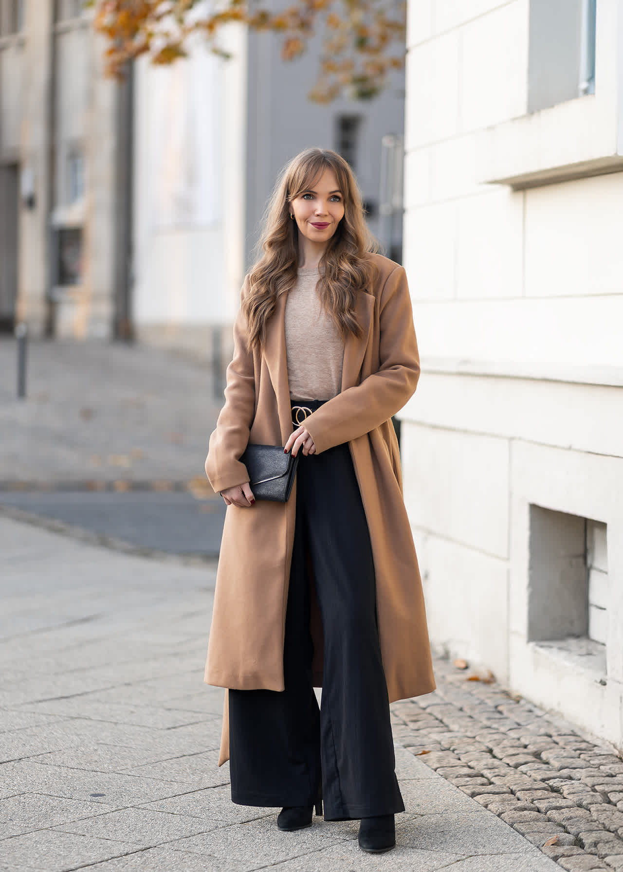 Frau in langem beigefarbenem Mantel, beigem Oberteil, weiten schwarzen Hosen, schwarzem Gürtel, schwarzer Clutch und schwarzen Stiefeletten.