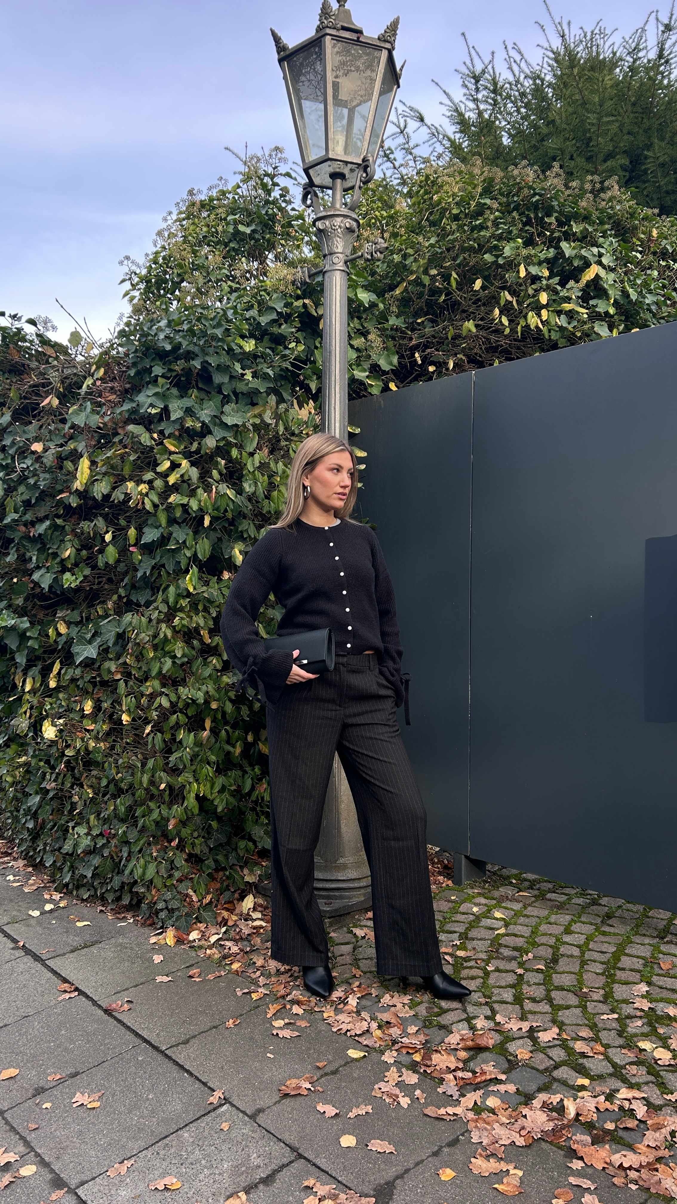Blonde Frau posiert vor Laterne, kombiniert schwarzen Cardigan mit passender Hose und Boots
