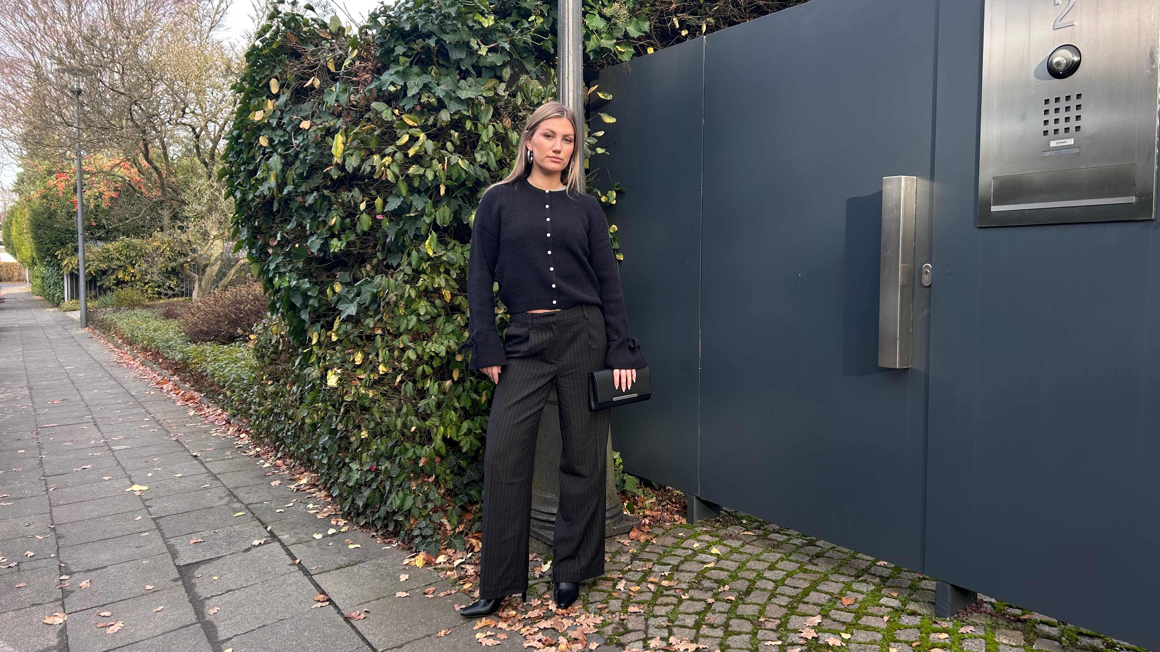 Junge Frau in schwarzem Cardigan und schwarzer Hose, steht vor Gartentor
