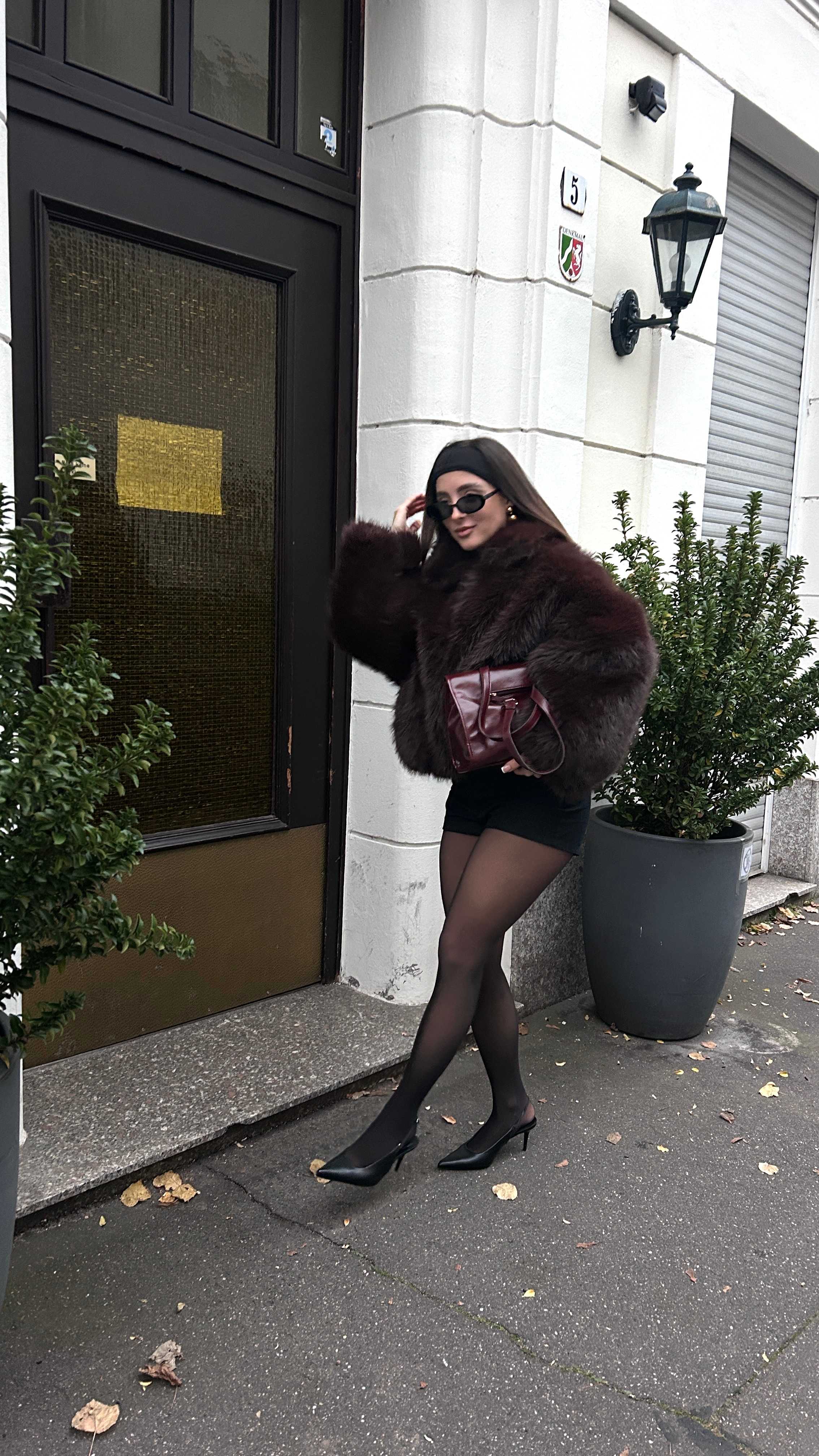 Frau in Mob Wife-Outfit, mit roter Clutch unterm Arm, steht in Hauseingang