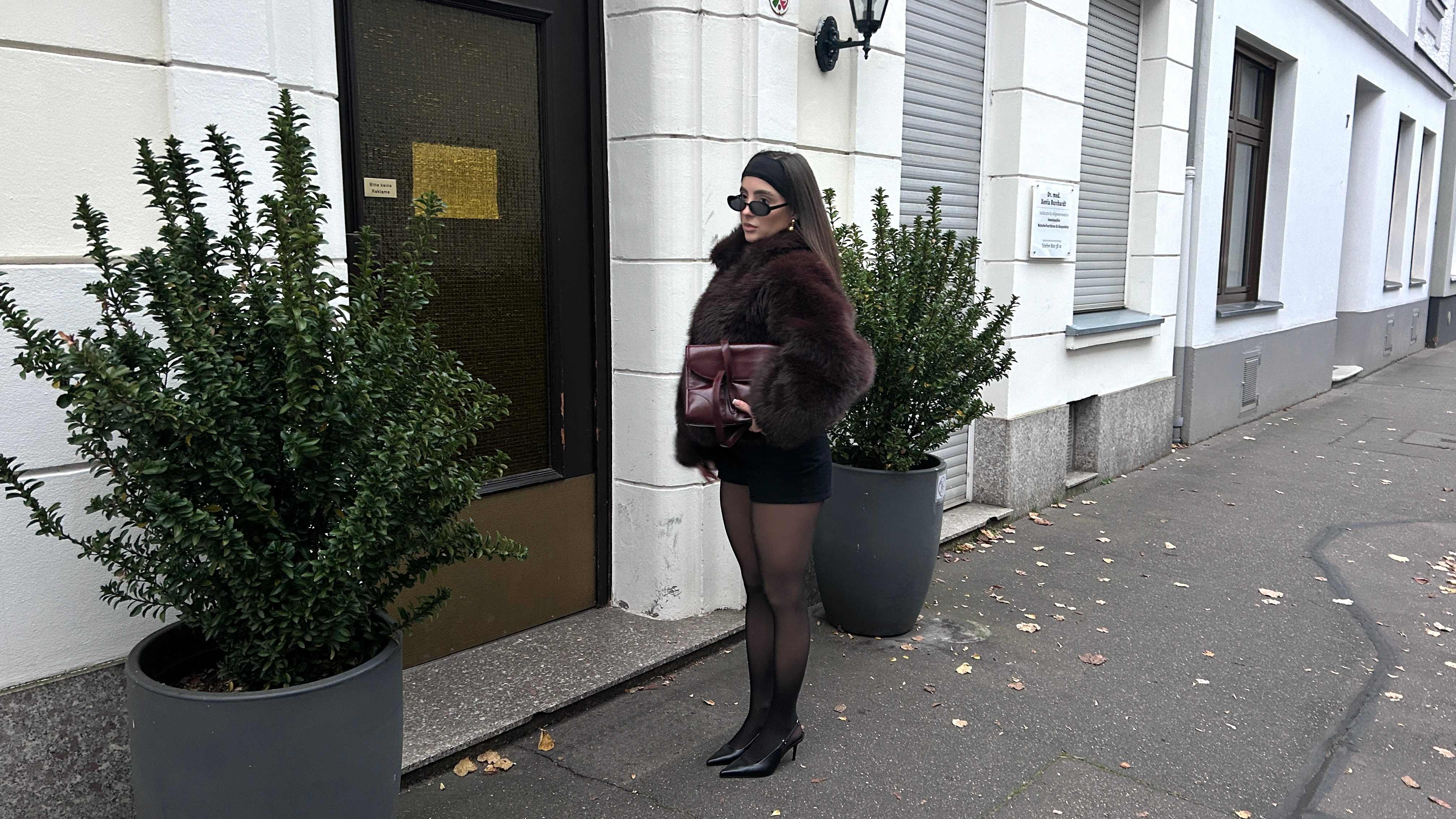 Frau trägt Outfit im Mob Wife-Style, kombiniert mit roter Handtasche, steht vor weißem Gebäude