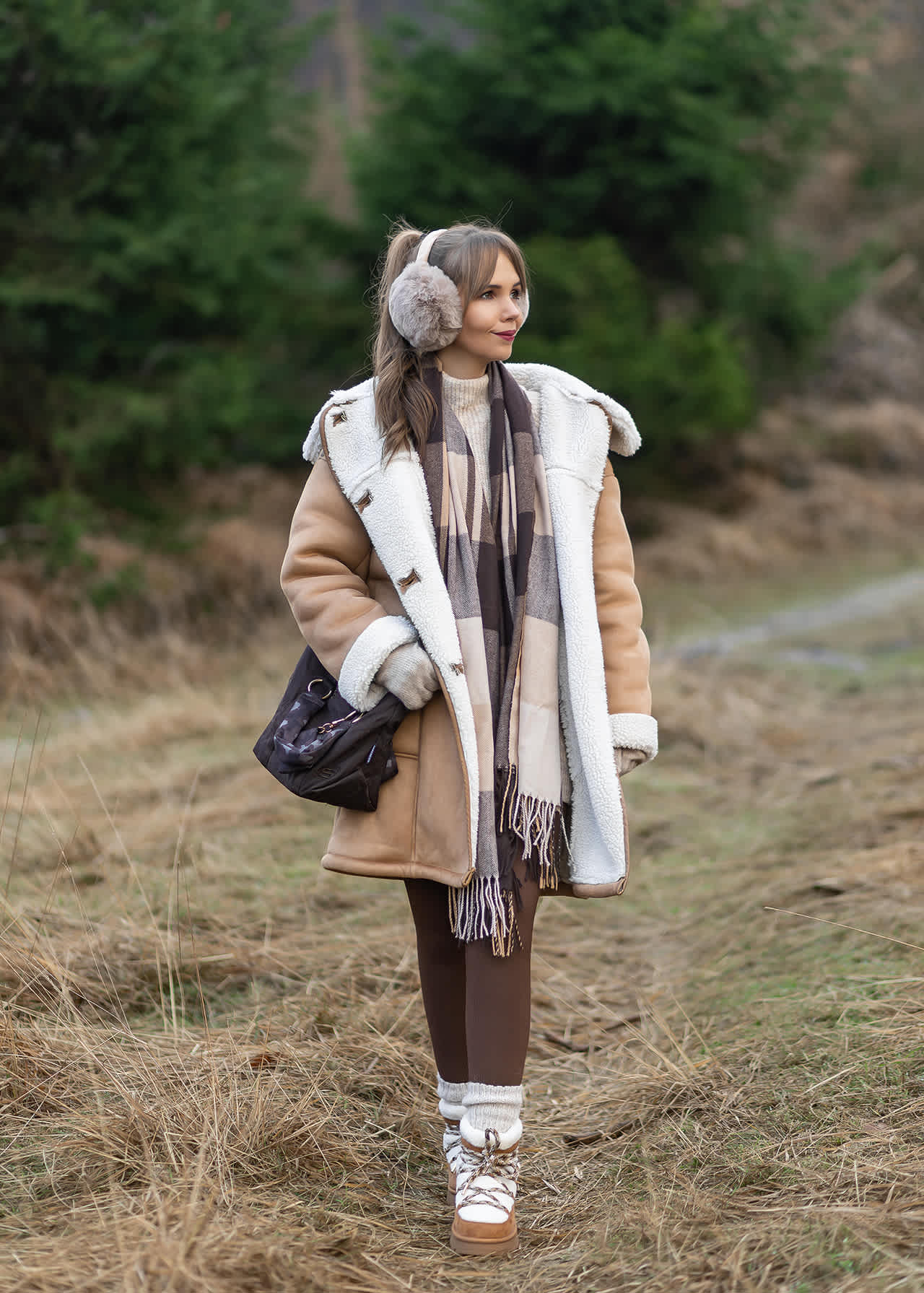 Frau in braun-weißem Outfit mit flauschigen Ohrenwärmern, steht auf Wiese