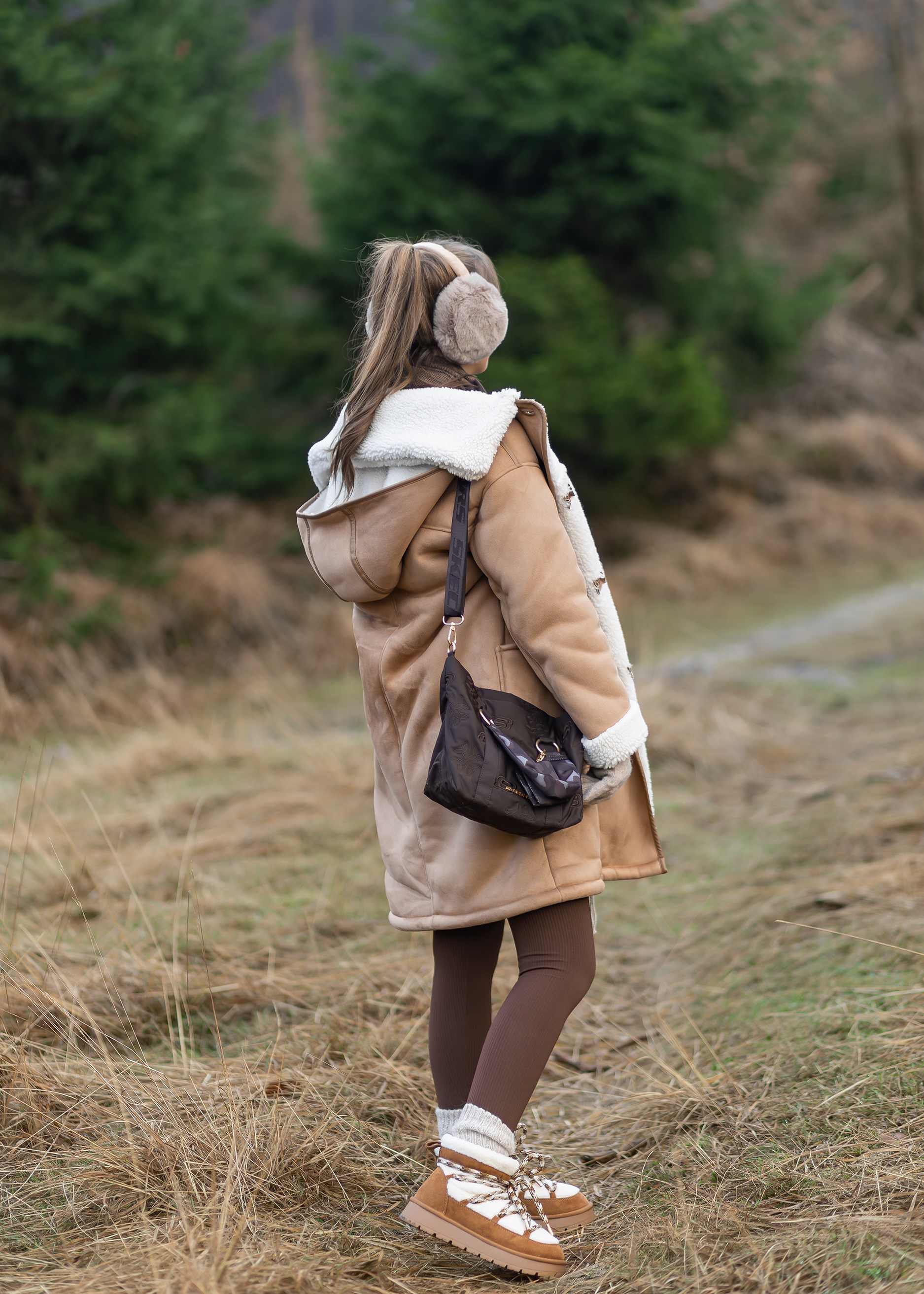 Frau in kuscheligem Kunstfell-Mantel, steht auf Wiese, trägt Handtasche und Ohrenwärmer in Flausch-Optik