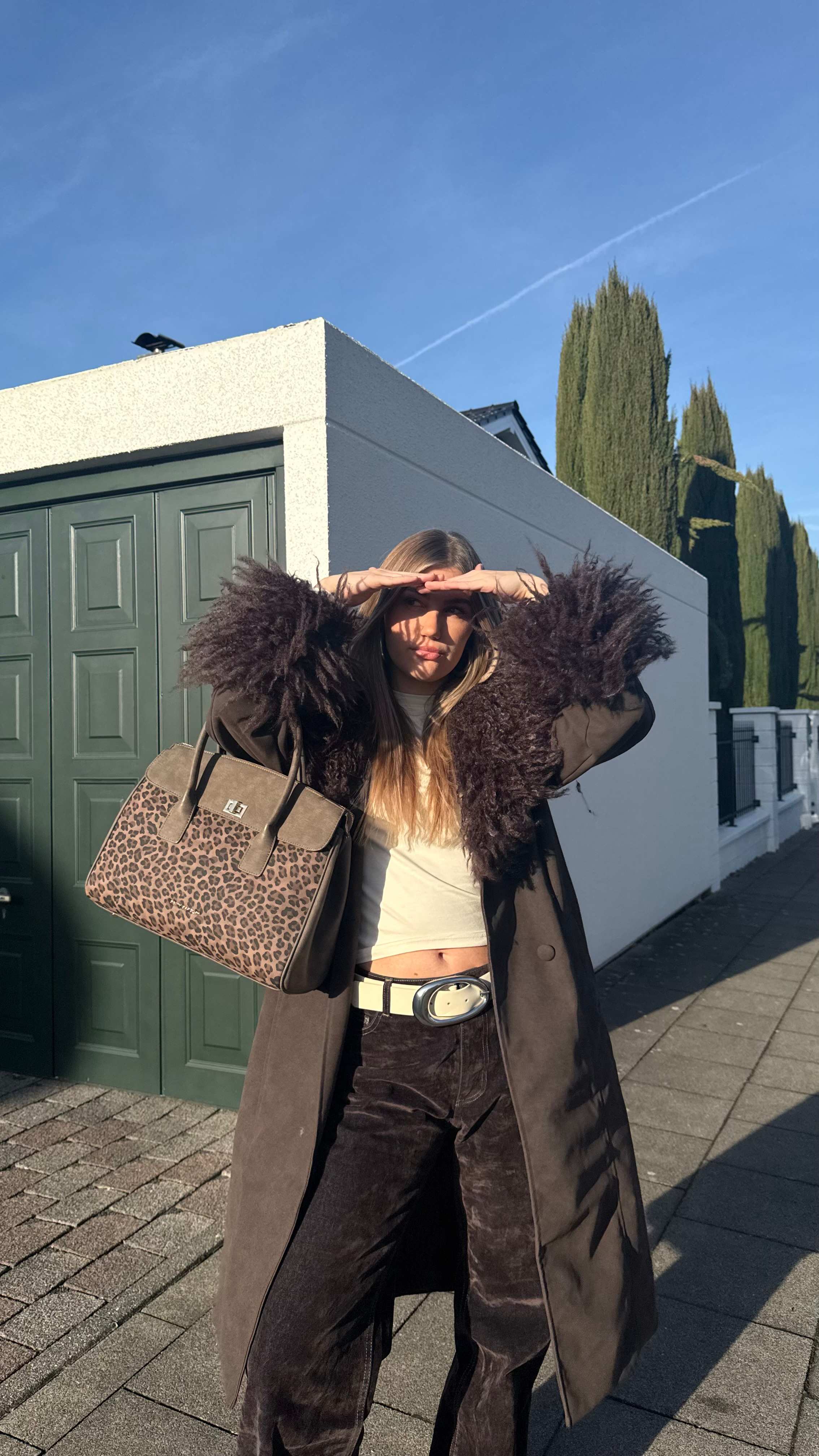Frau steht vor Garage, schützt ihre Augen vor der Sonne, trägt Animalprint-Tasche und Felljacke