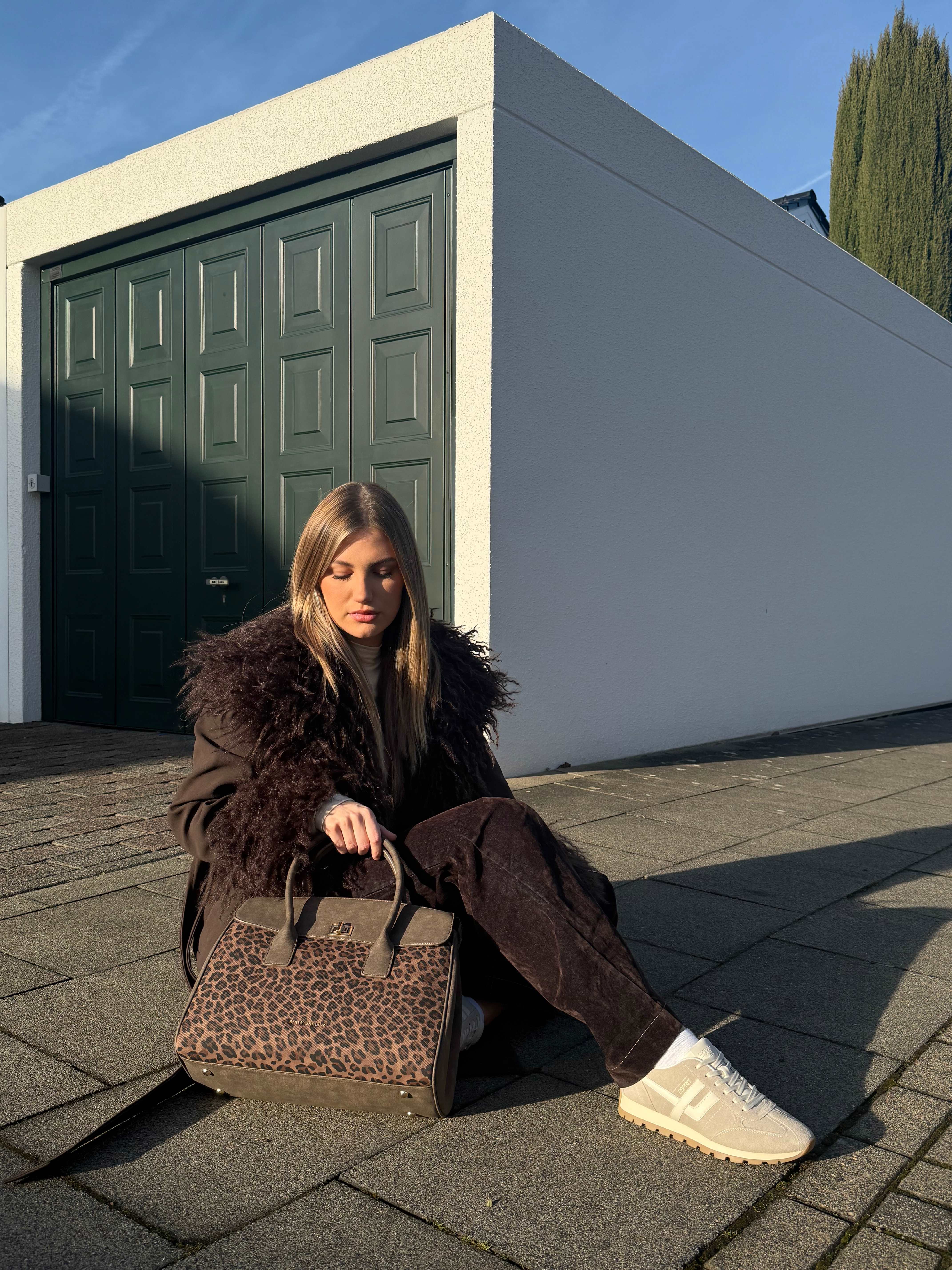 Blonde Frau sitzt auf Boden vor Garage, mit Animalprint-Handtasche