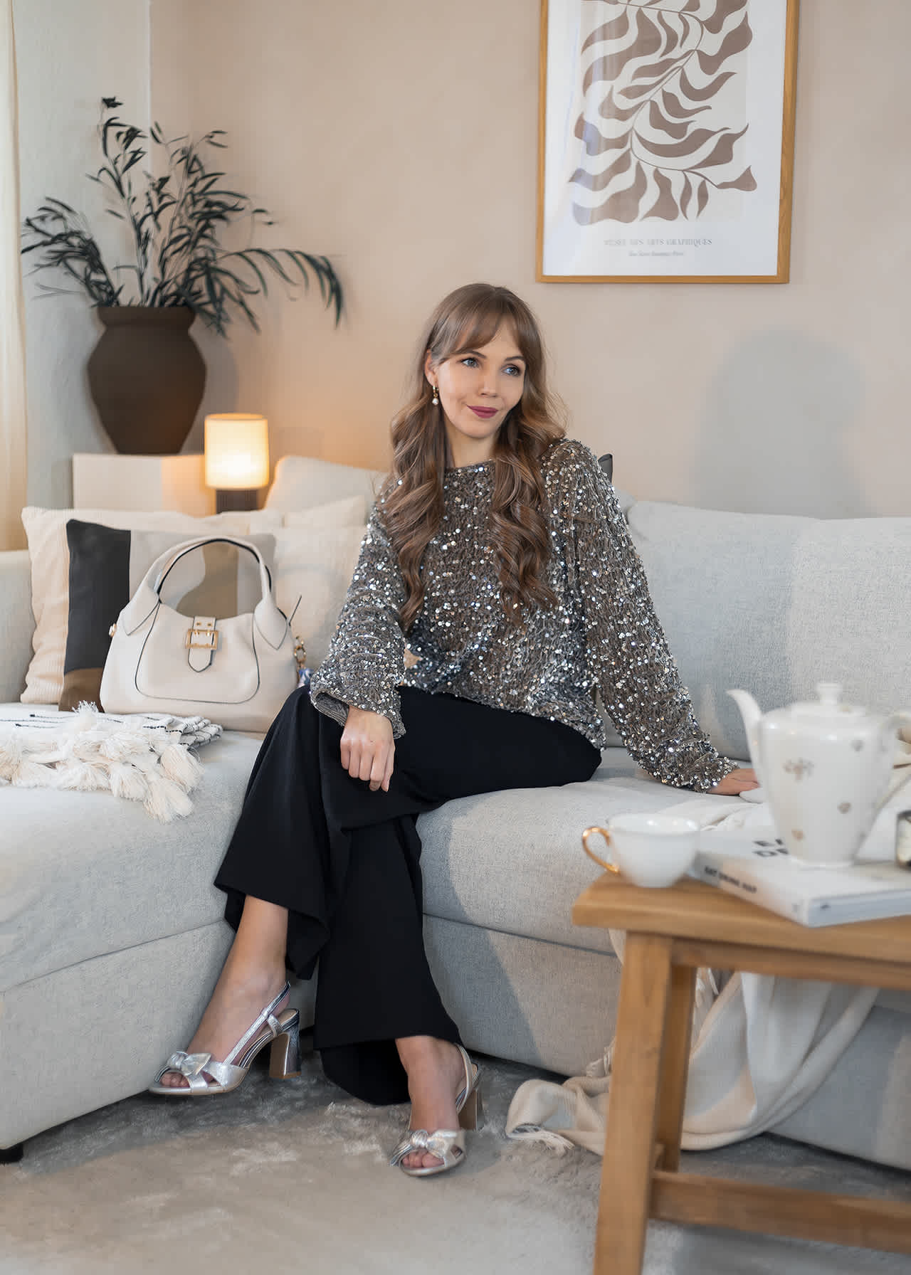 Frau sitzt auf Sofa, trägt schwarze Hose mit Glitzer-Oberteil und silbernen Bridgerton-Sandaletten
