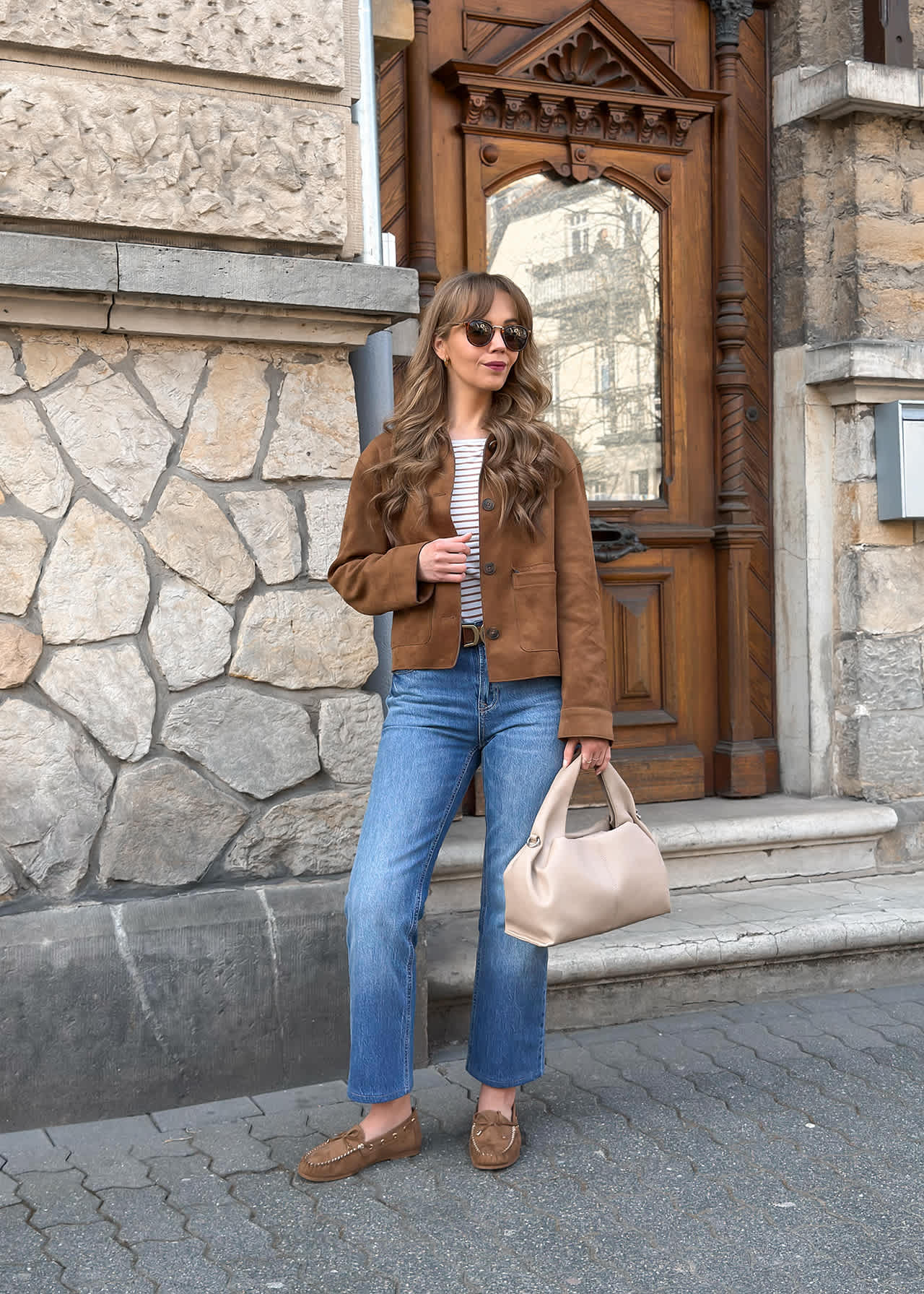 Frau steht vor einer Holztür, trägt eine braune Wildlederjacke zu einer blauen Jeans und passenden braunen Mokassins 