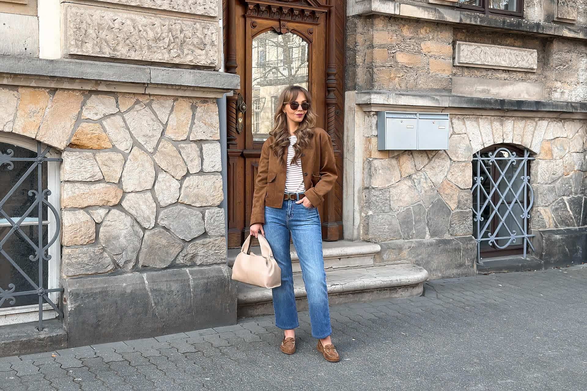 Frau präsentiert Outfit vor einer Holztür, trägt braune Lederjacke zu gestreiftem Shirt, Jeans und passenden Mokassins.