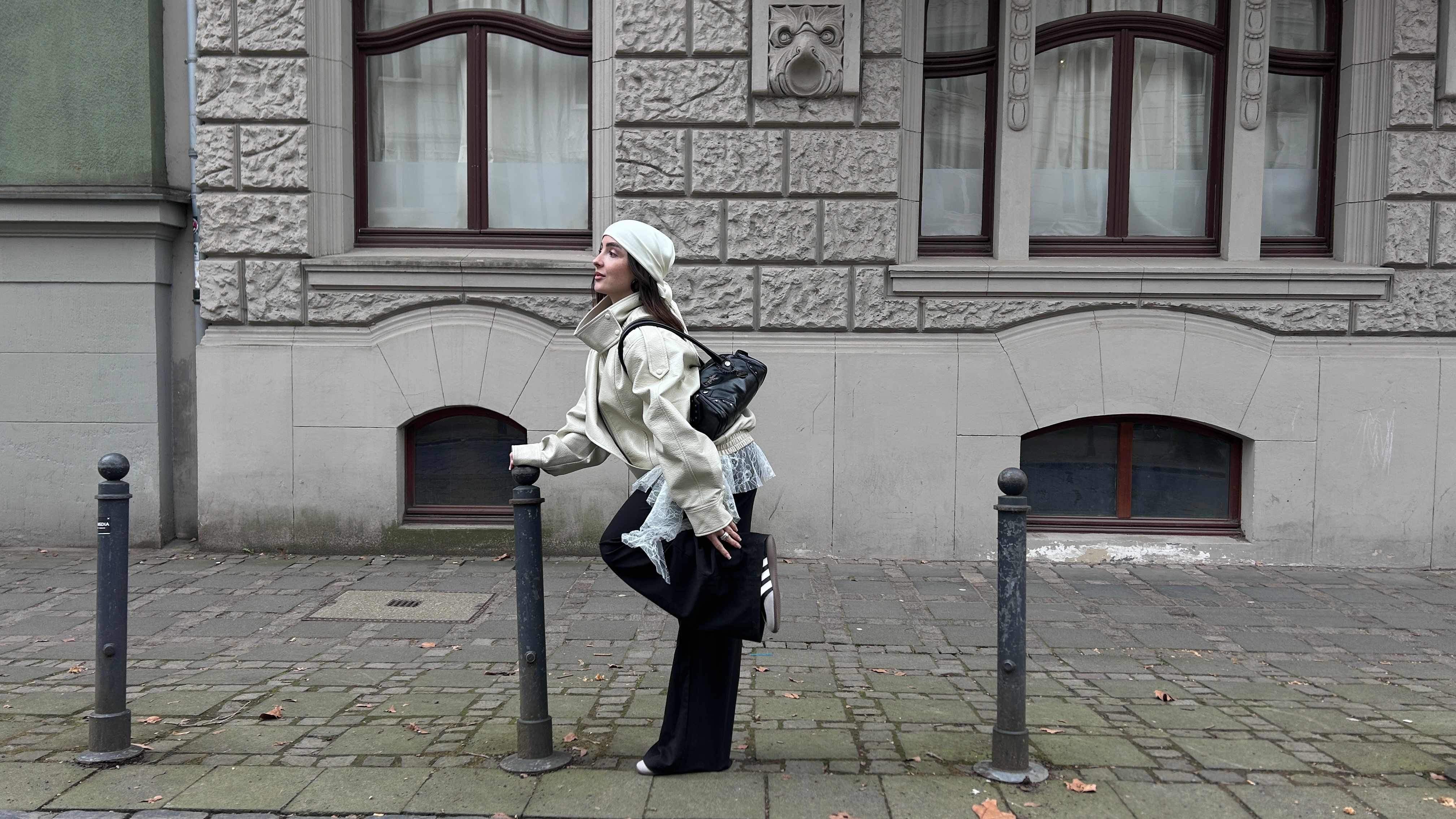 Eine Frau zeigt ihr Outfit. Fokus auf den sportlichen schwarz-weißen Riemchen-Ballerinas. Urbaner Look in Creme und Schwarz mit Oversize-Lederjacke, Mesh-Detail und Kopftuch.