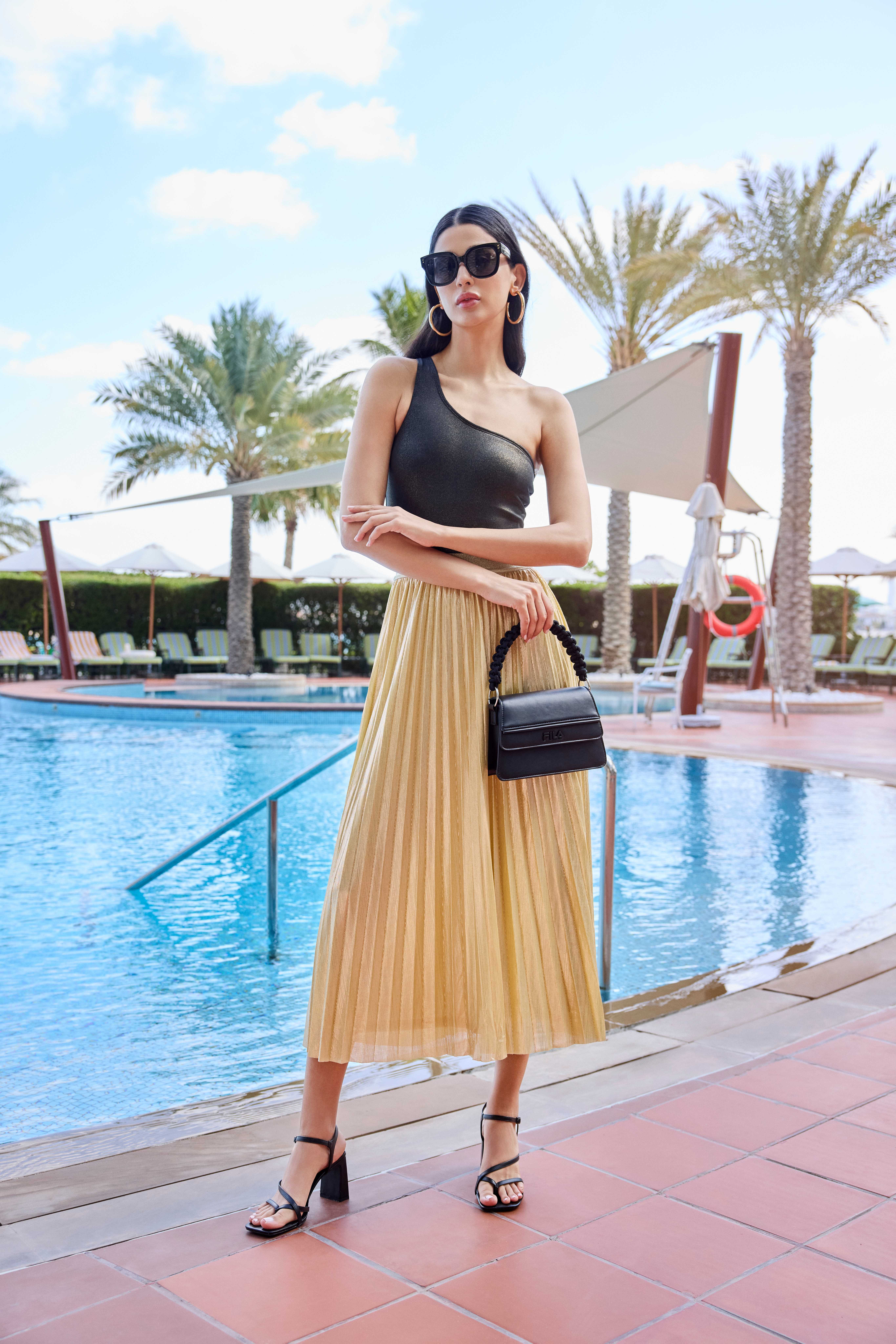 Schwarzhaarige Frau trägt eleganten Oneshoulder-Badeanzug kombiniert mit langem Plisseerock, stehend vor Pool