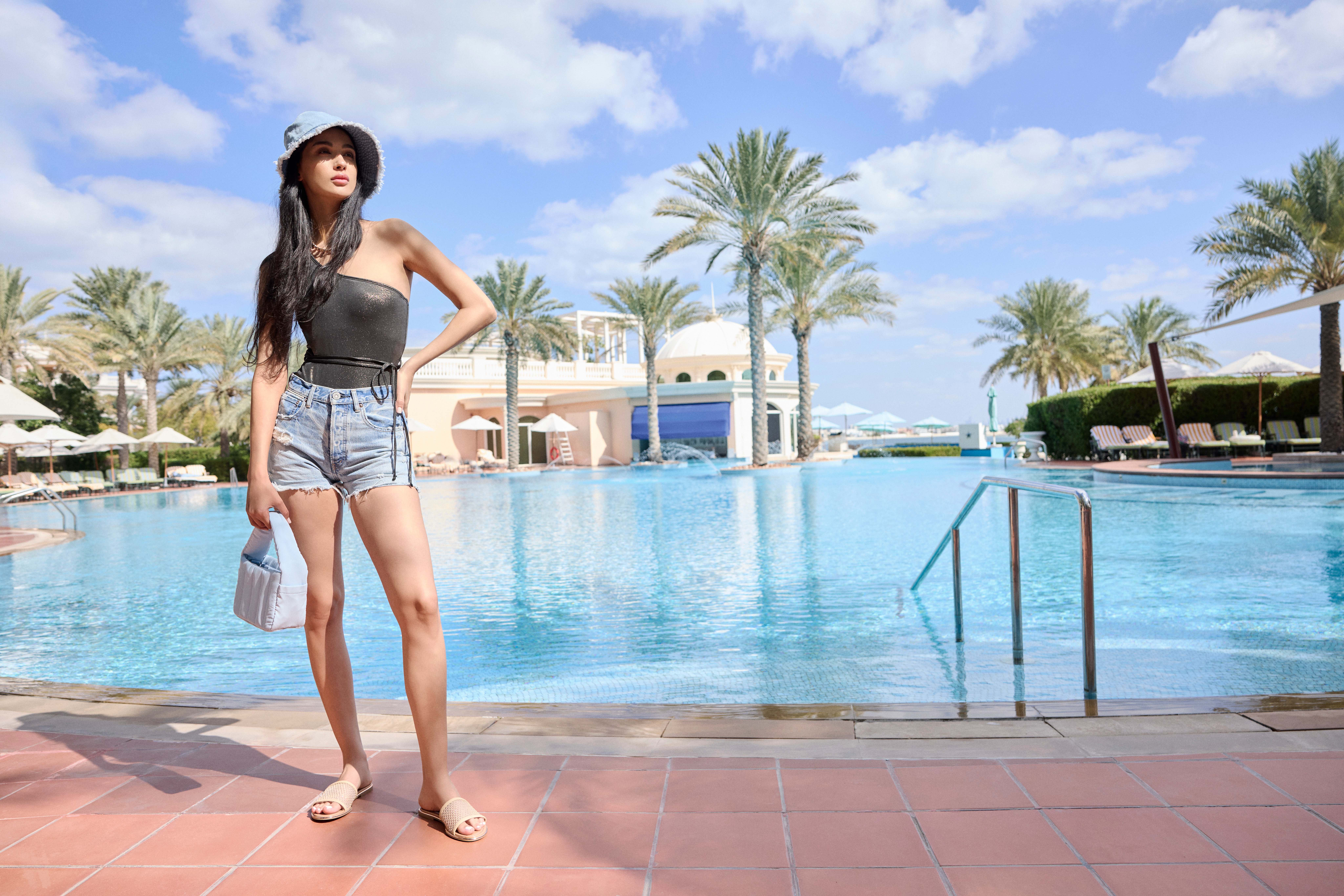 Frau mit grünem Top, Denim-Pants und Bucket Hat, steht direkt vor großem Swimming Pool