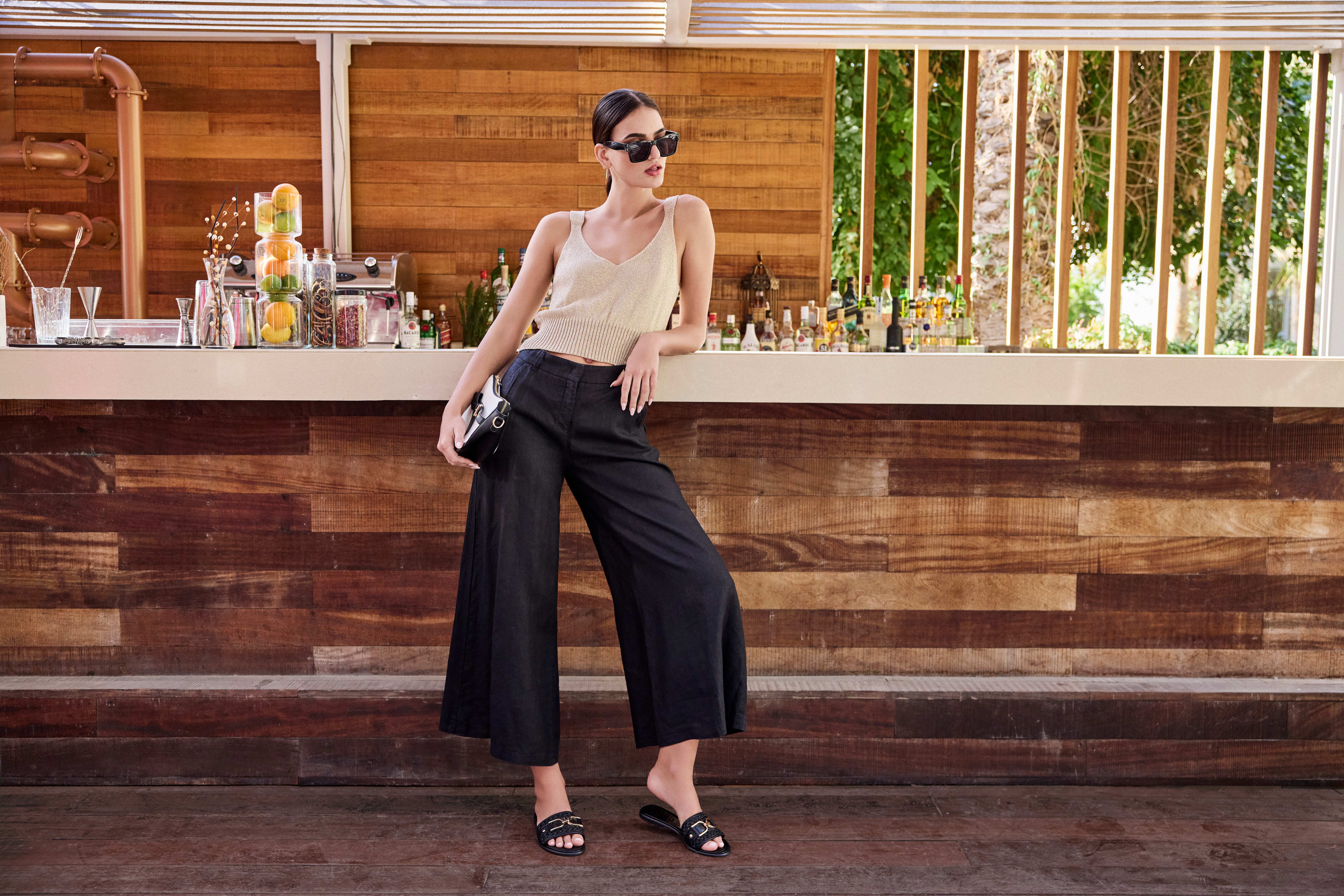 Frau mit elegantem Top, schwarzer Hose und Clutch, trägt Sonnenbrille und lehnt an Bar