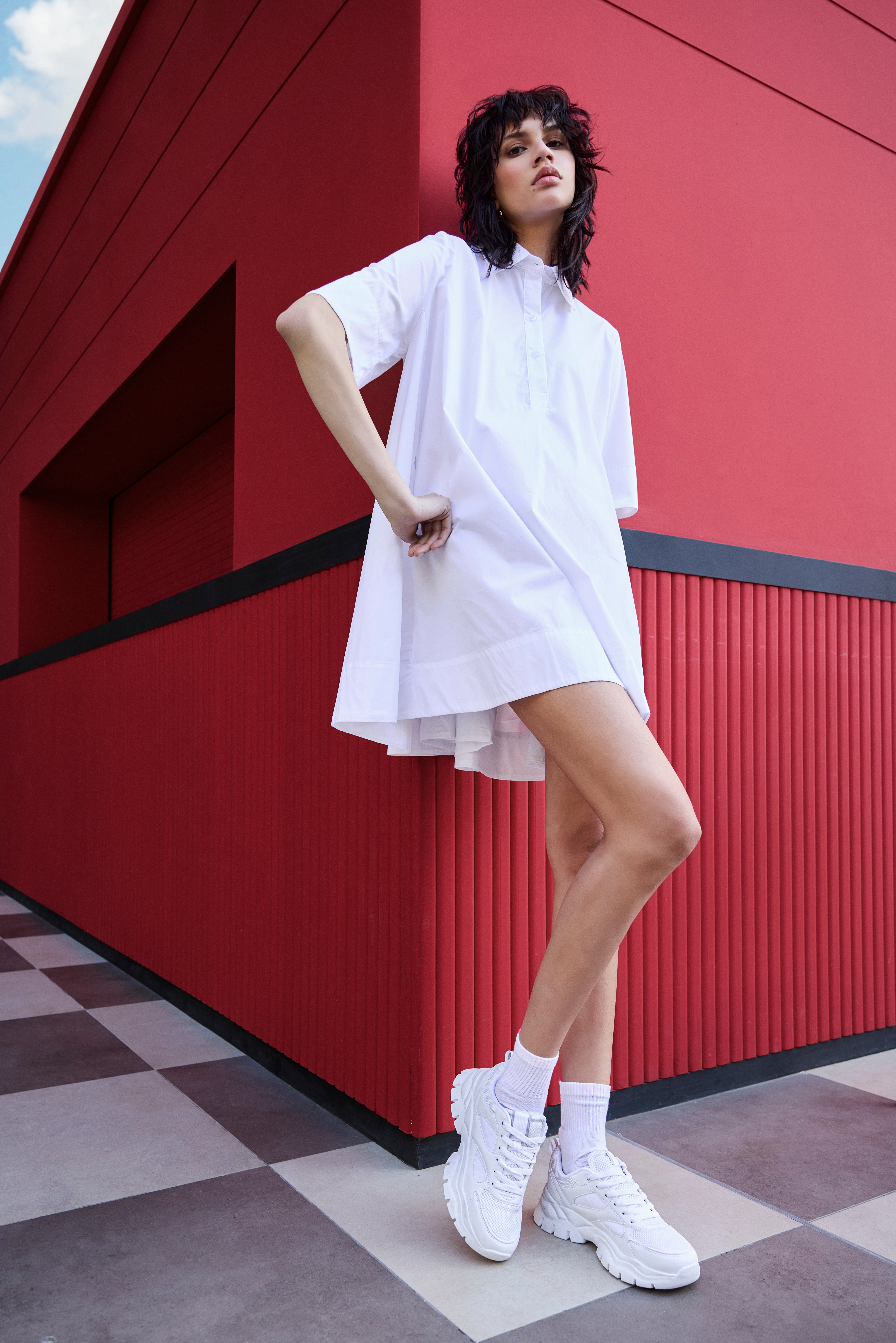 Frau in Oversized Shirt und weißen Shorts im All White Outfit, stehend an der Ecke eines roten Baus
