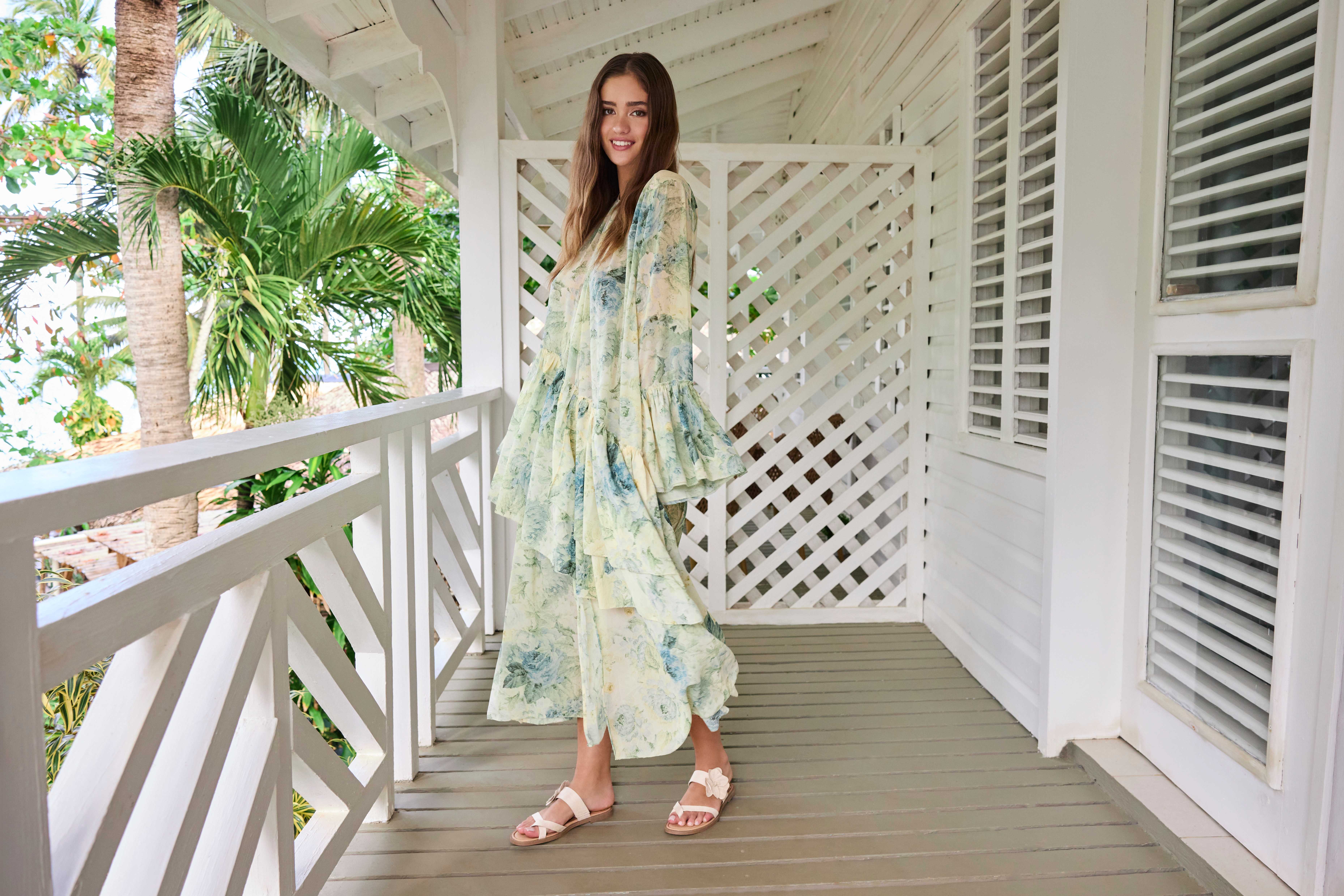 Lächelnde Frau in locker-fallendem Blumenkleid und Sandalen, steht auf Veranda