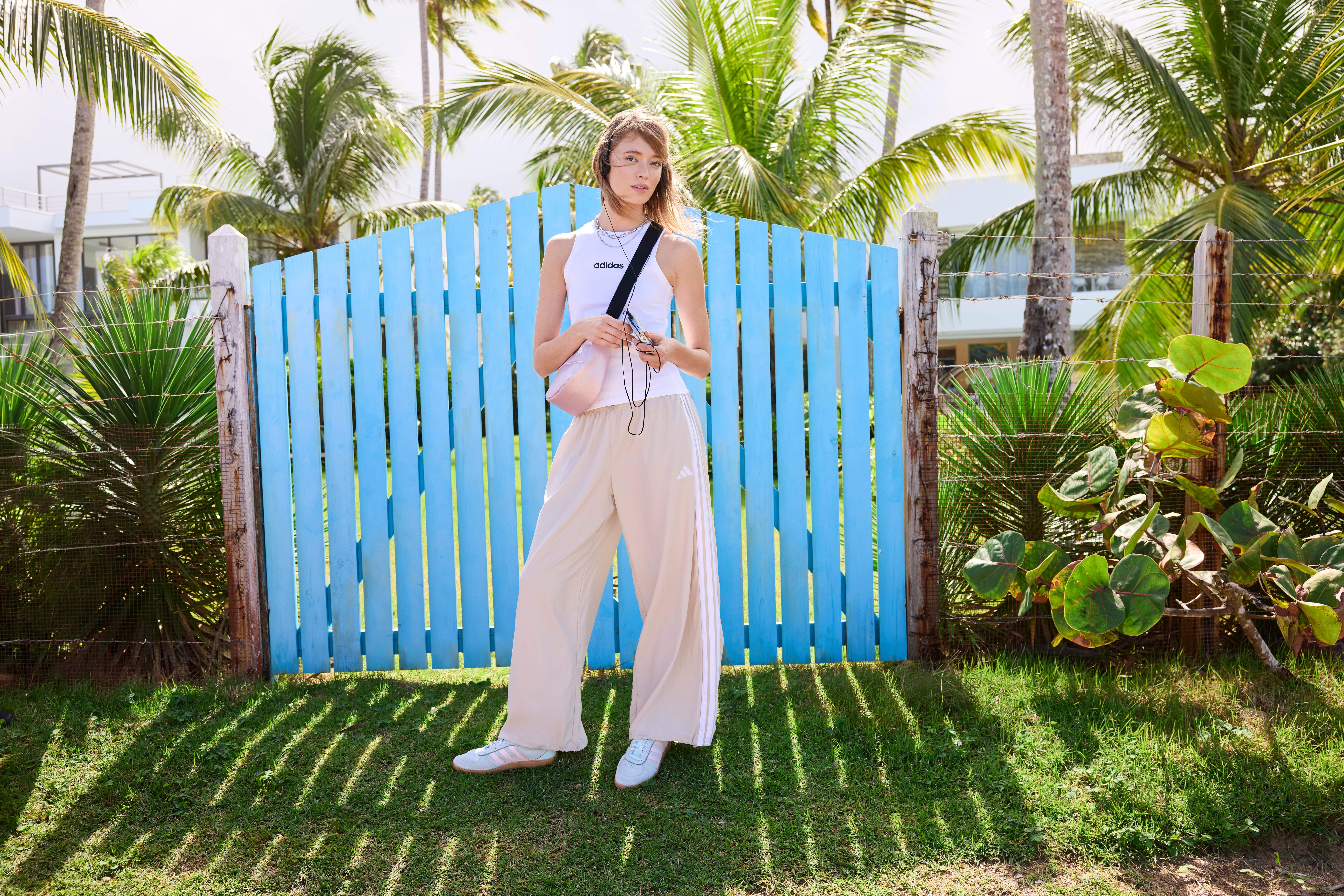 Frau in weißem Top und beigen Pants, steht vor blauem Gartentor, in Palmenkulisse
