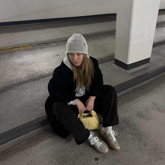 Frau sitzt auf Boden von Betonrampe, trägt Metallic Sneaker, dazu schwarzen Mantel