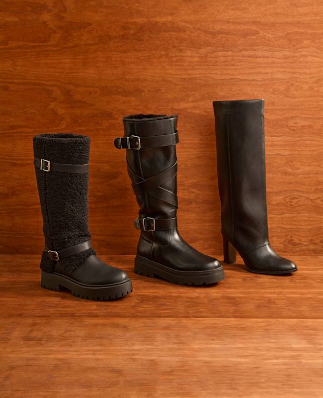Drei schwarze Lederstiefel mit weitem Schaft vor Holzpaneel