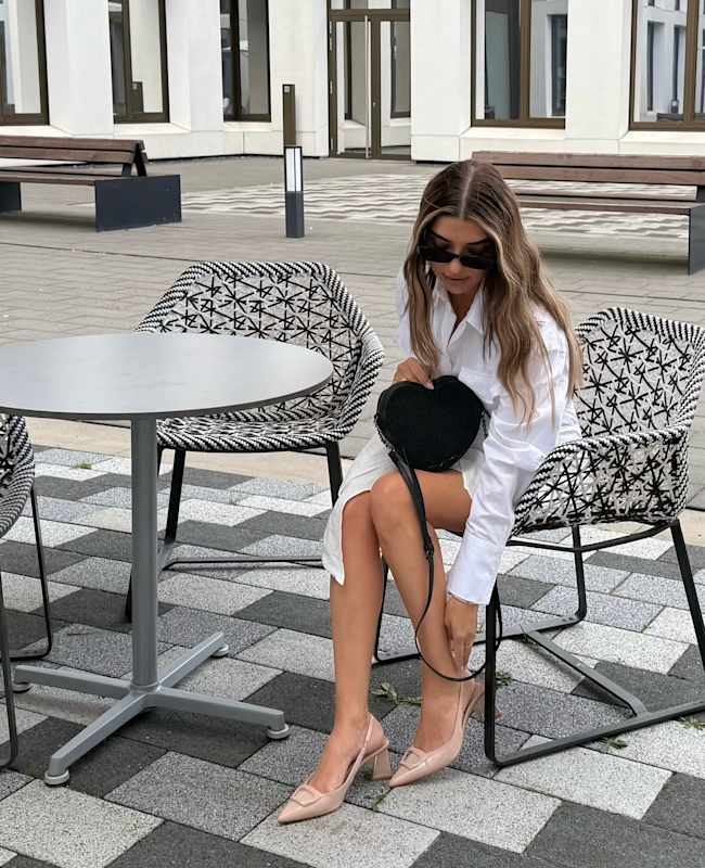 Junge Frau sitzt in weißem Outfit mit schwarzer Glitzertasche an Außentisch im Stadtbereich