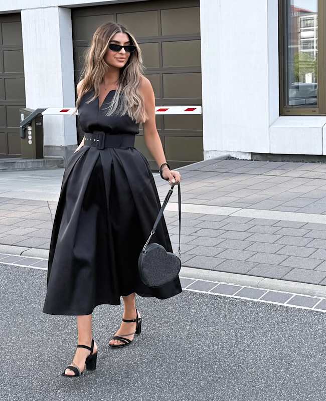 Junge Frau in elegant-schwarzem Kleid und Sonnenbrille, kombiniert mit schwarzer Glitzertasche in Herzform