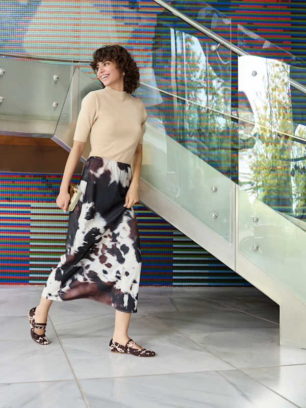 Frau in braunem Top mit langem Cow-Print-Rock, stehend vor Glastreppe