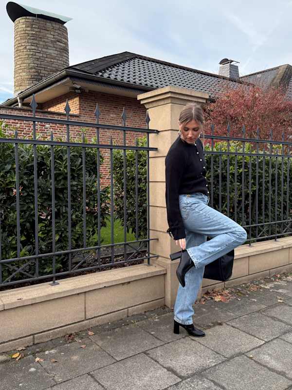 Frau trägt schwarzen Cardigan in Kombination mit blauer Jeanshose, lehnt an steinerner Einzäunung